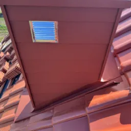 Czerwona dachówka ceramiczna z zamontowanym wywietrznikiem dachowym z metalową kratką wentylacyjną, widok z bliska na fragment dachu.