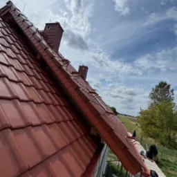 Czerwona dachówka ceramiczna na ukończonym dachu z kominem, widoczna z rusztowania na tle błękitnego nieba z chmurami i zielenią w oddali.