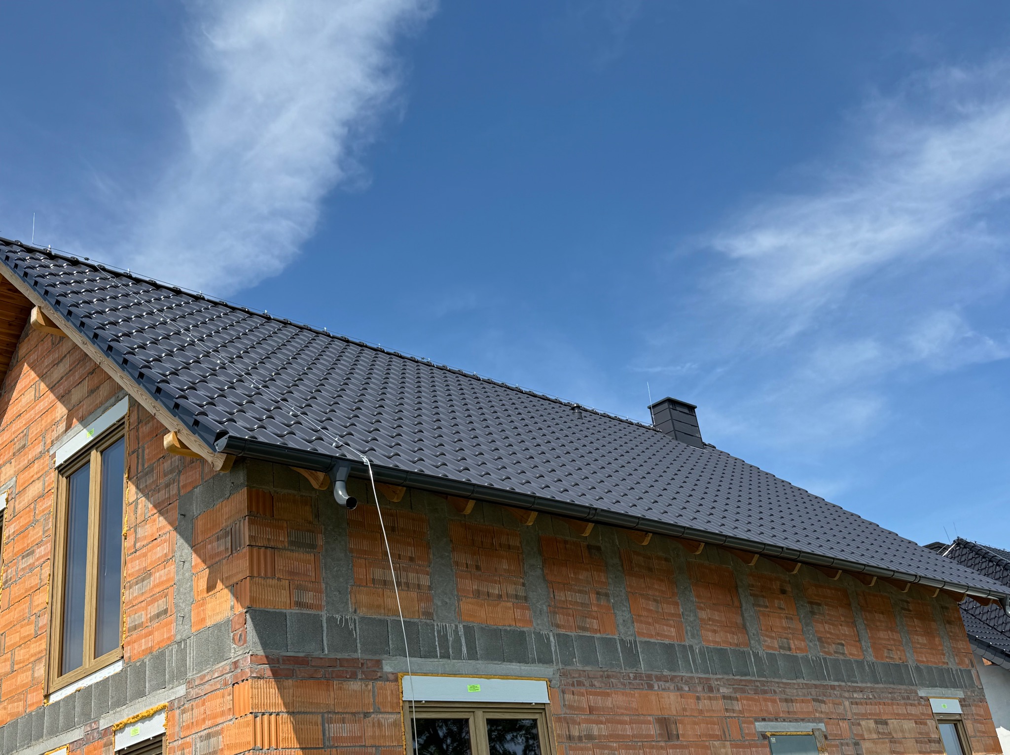 Nowy, błyszczący dach z ciemnej dachówki ceramicznej na budynku w trakcie budowy, widoczne rynny i fragment komina na tle błękitnego nieba.