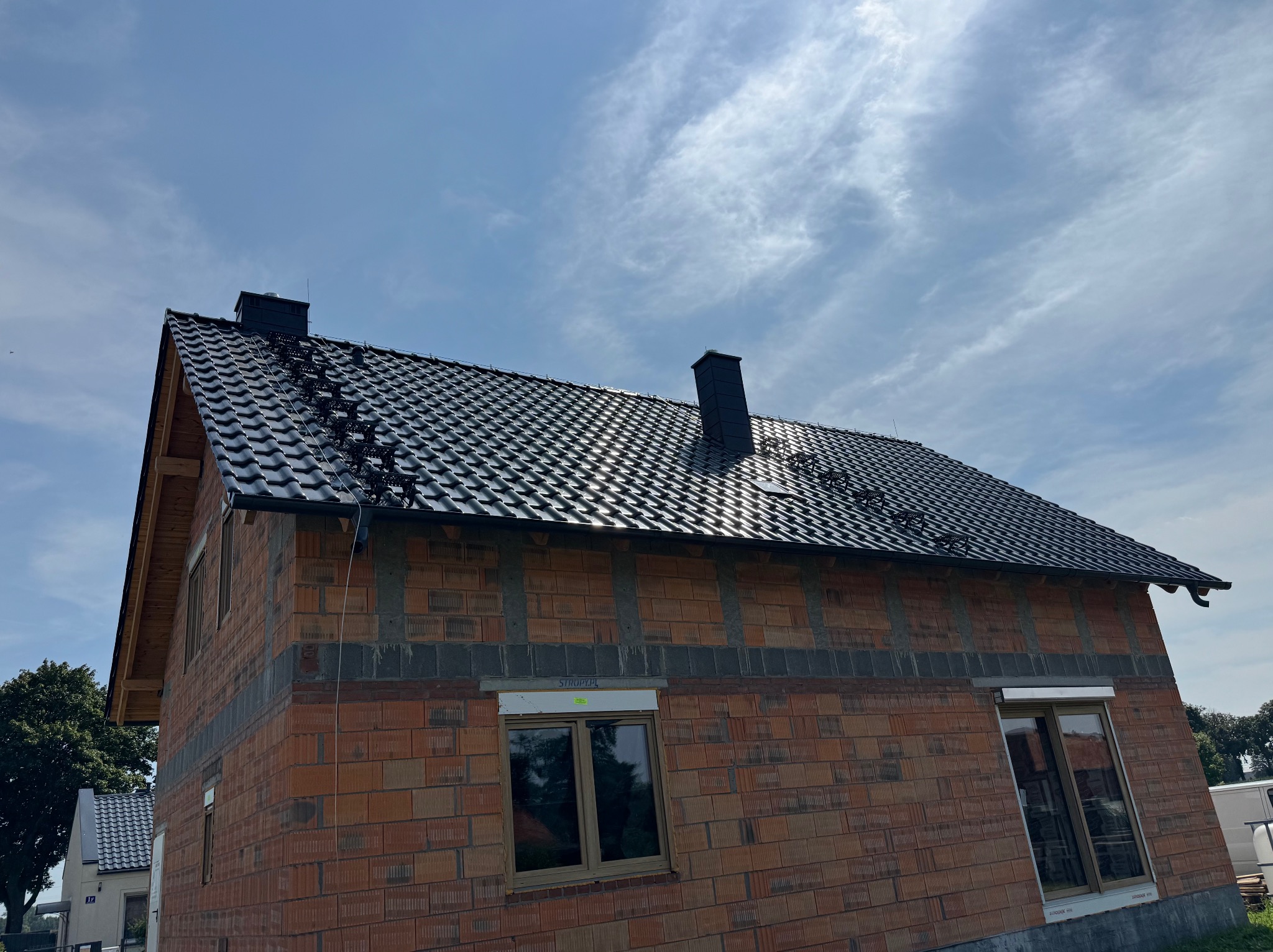 Ceglany dom w budowie z czarnym dachem z dachówki ceramicznej i zamontowanymi rynnami. Widoczne kominy i fragment nieba z chmurami.
