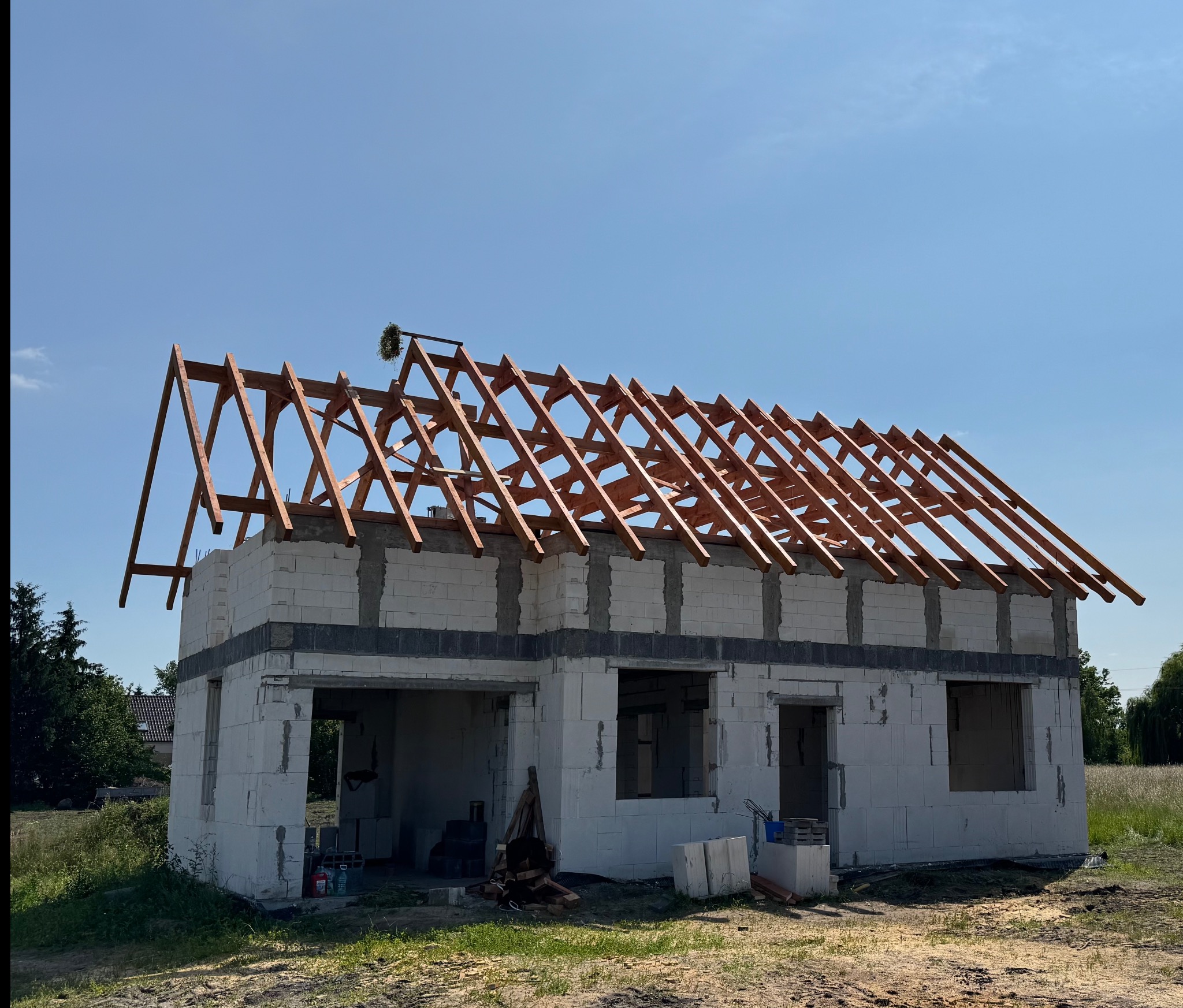 Wznoszenie więźby dachowej na budynku z białych bloczków, widoczne elementy konstrukcyjne z drewna, wieniec na dachu, błękitne niebo w tle.