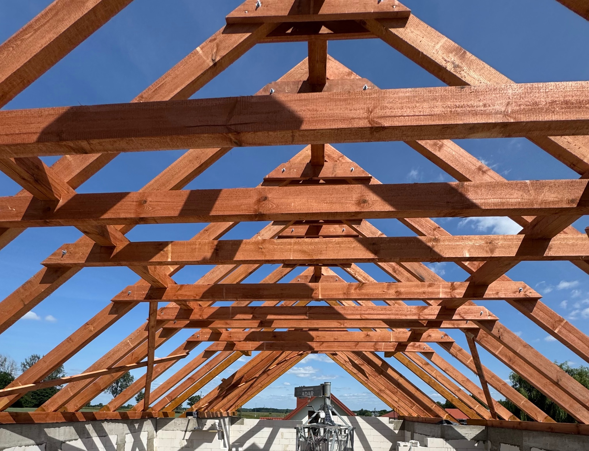 Wnętrze konstrukcji dachu z brązowych belek pod błękitnym niebem, widok z dołu, elementy łączone śrubami, w tle krajobraz z domami i zielenią.
