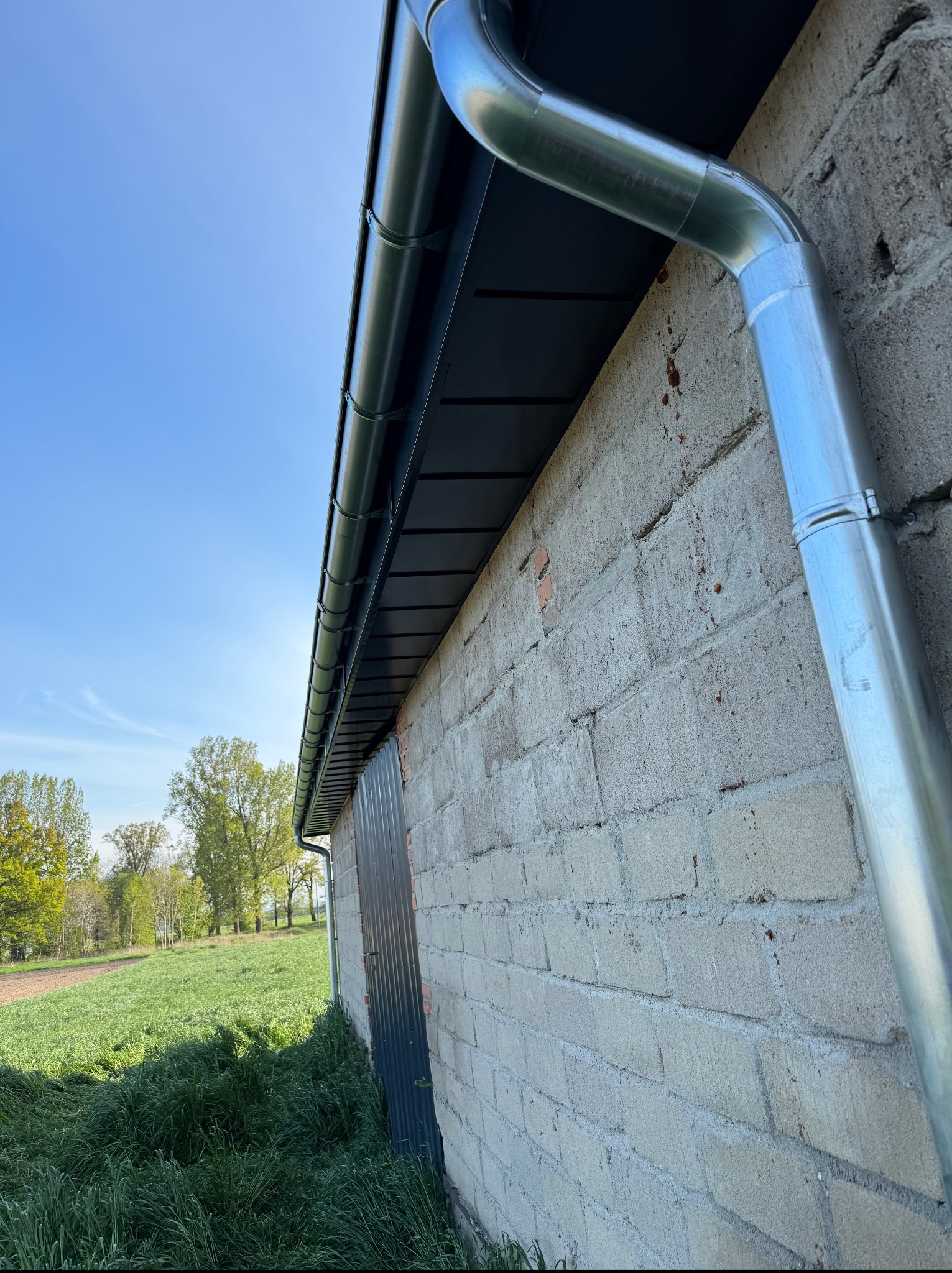 Zamontowana, srebrna rynna pionowa i pozioma na szarym murze budynku z widokiem na zielone pole i drzewa w tle. Ujęcie z dołu, w słoneczny dzień.