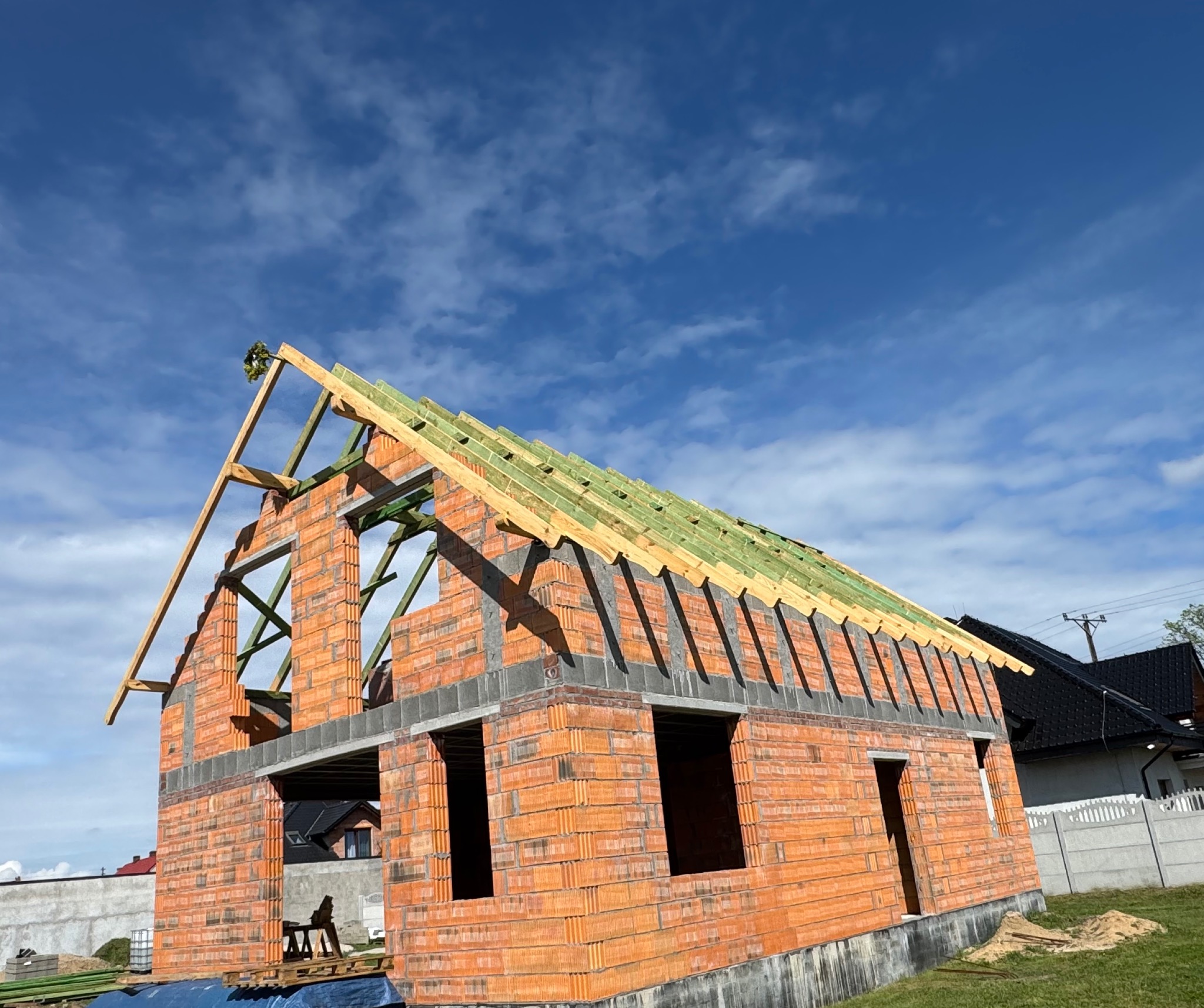 Niedokończony budynek z cegły z widoczną konstrukcją dachu z drewnianych belek i zielonych łat, na tle błękitnego nieba z chmurami.
