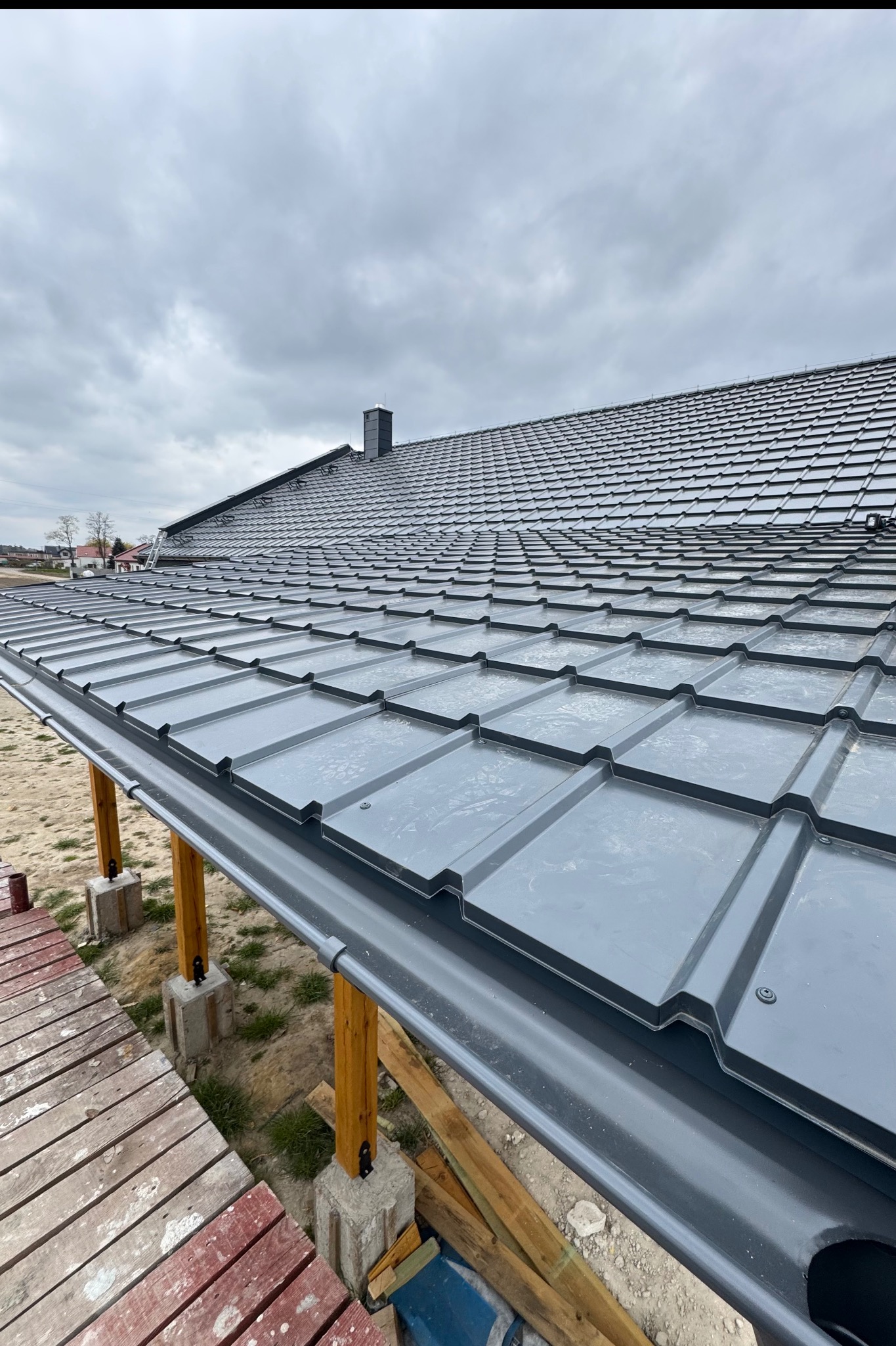 Szare dachówki na nowym dachu domu z zamontowanymi rynnami, widziane z perspektywy rusztowania. Pochmurne niebo w tle.