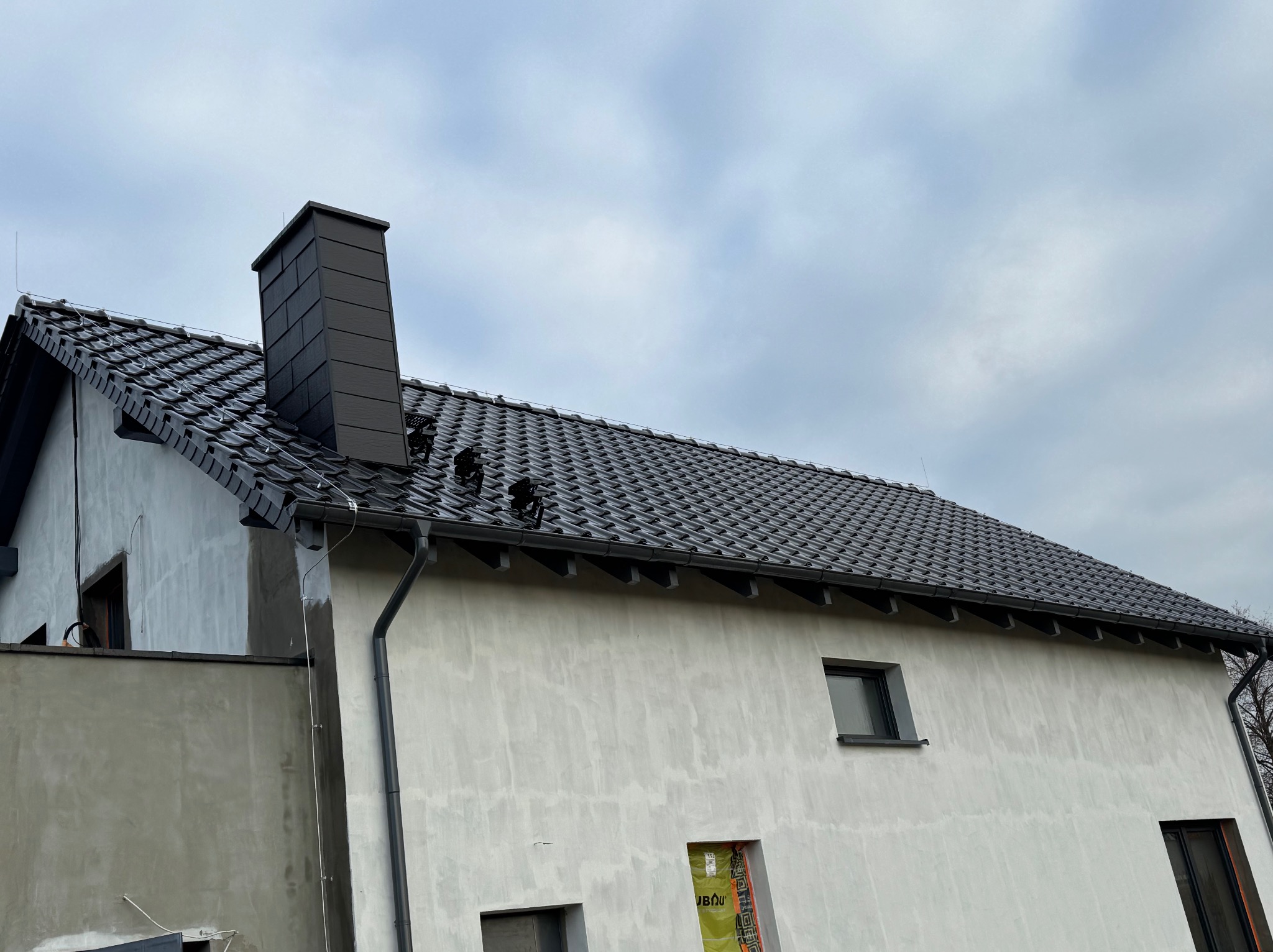 Dom w trakcie budowy z nowym dachem z czarnej dachówki karpiówki i czarną rynną. Widoczny komin i surowa elewacja. Pochmurne niebo w tle.