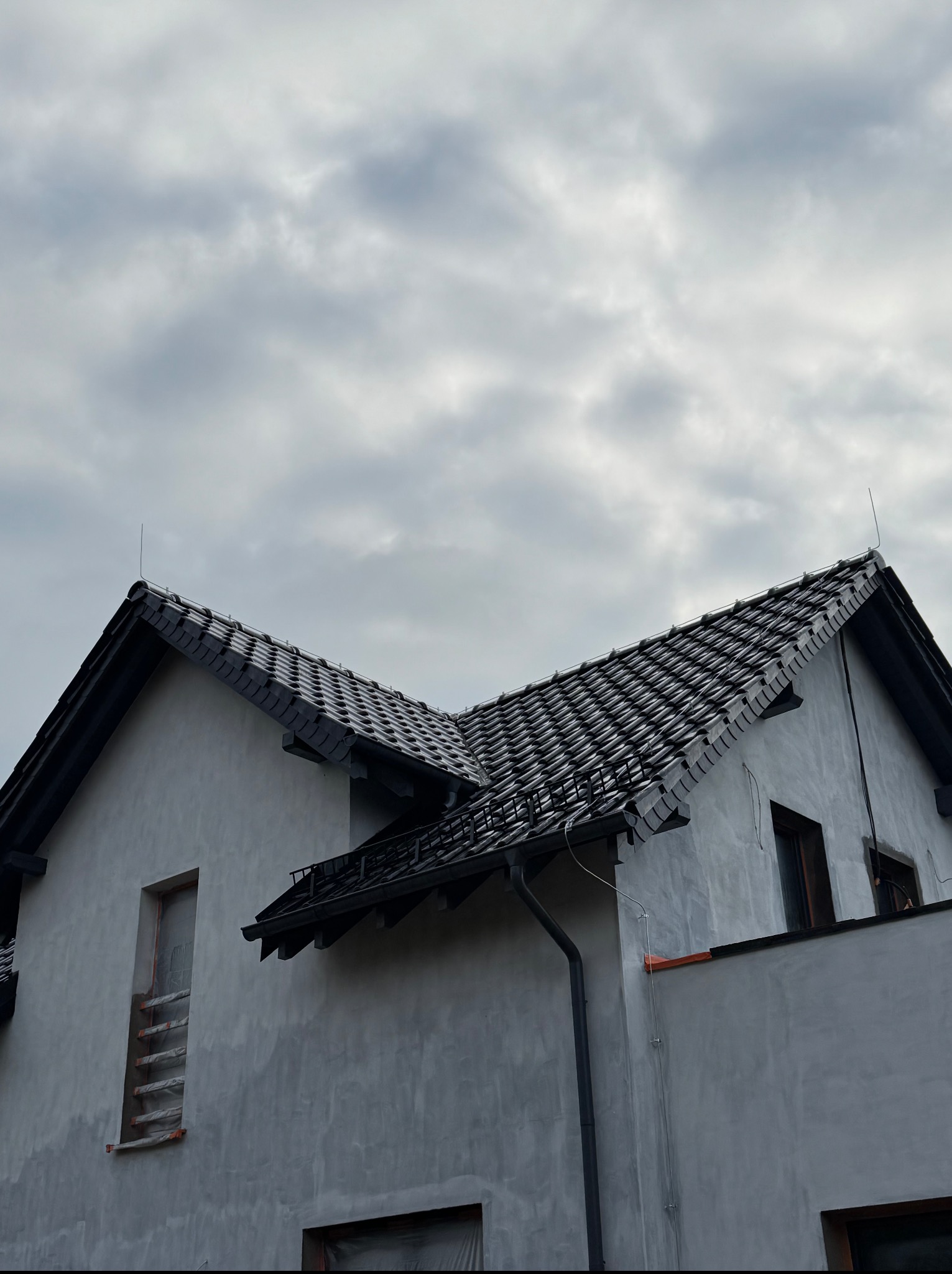 Dom jednorodzinny z nowym, ciemnym dachem z dachówki ceramicznej i rynnami. Widoczne zabezpieczone okna i elewacja w trakcie prac wykończeniowych.