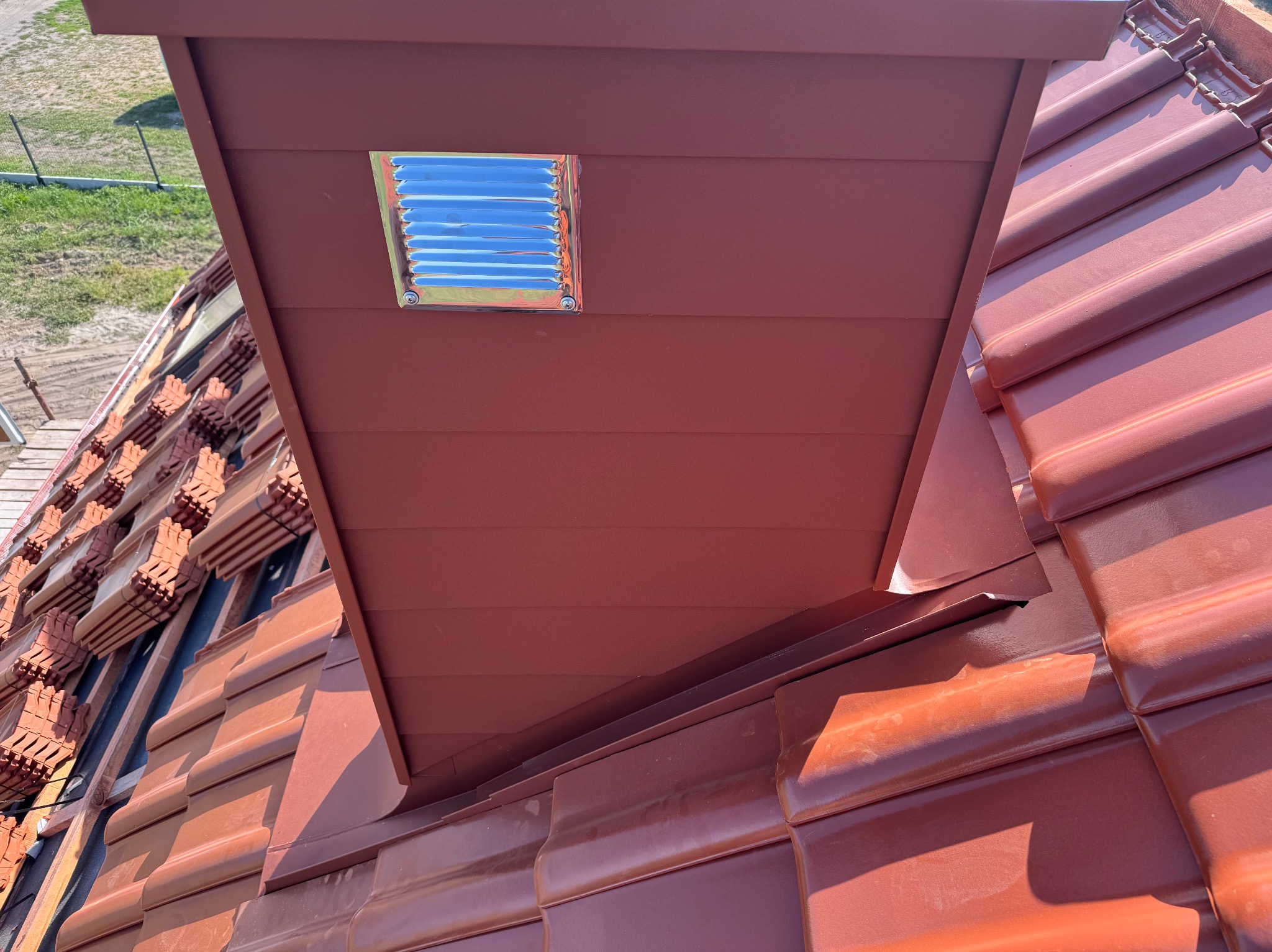 Czerwona dachówka ceramiczna z zamontowanym wywietrznikiem dachowym z metalową kratką wentylacyjną, widok z bliska na fragment dachu.