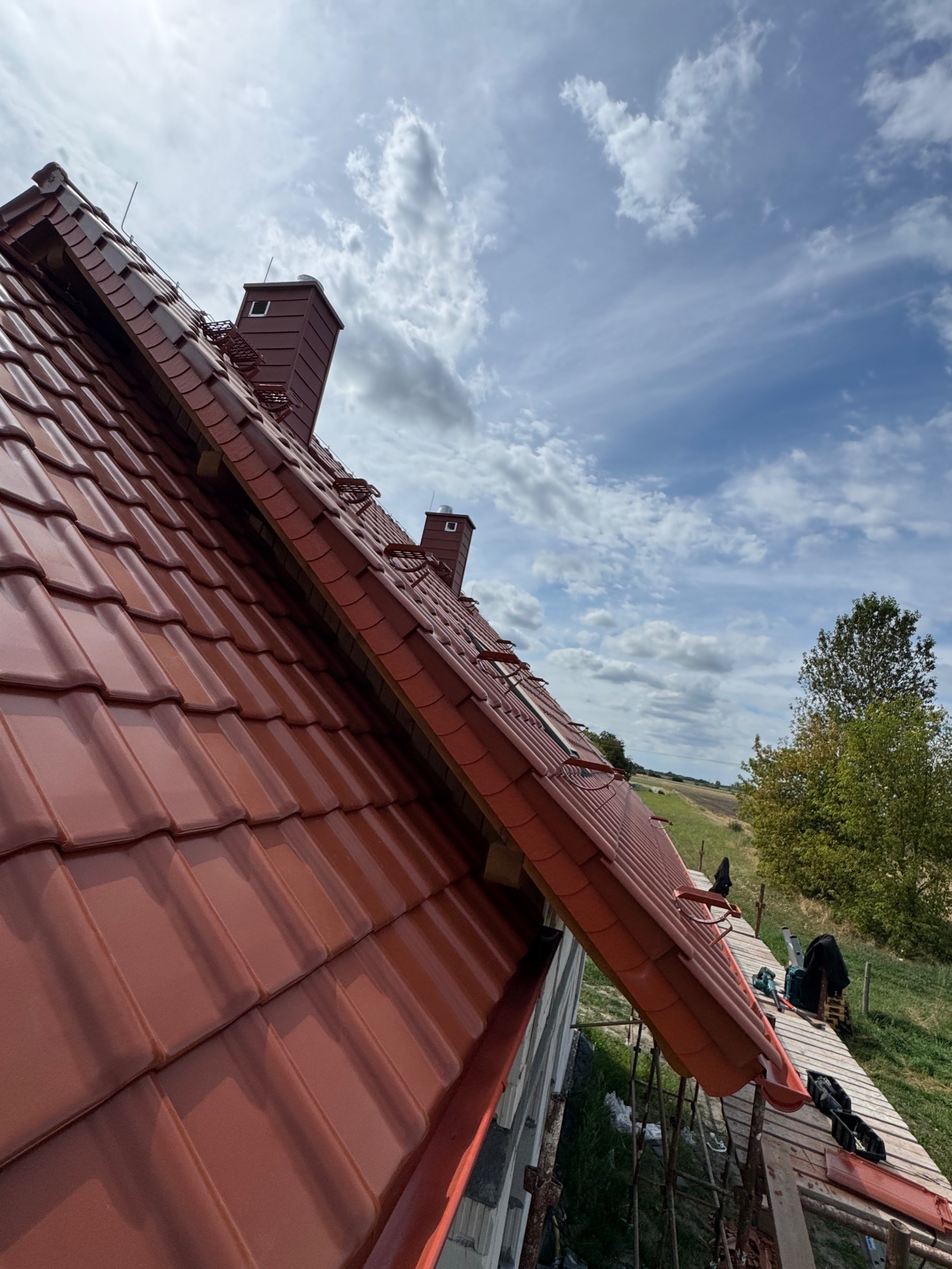 Czerwona dachówka ceramiczna na ukończonym dachu z kominem, widoczna z rusztowania na tle błękitnego nieba z chmurami i zielenią w oddali.