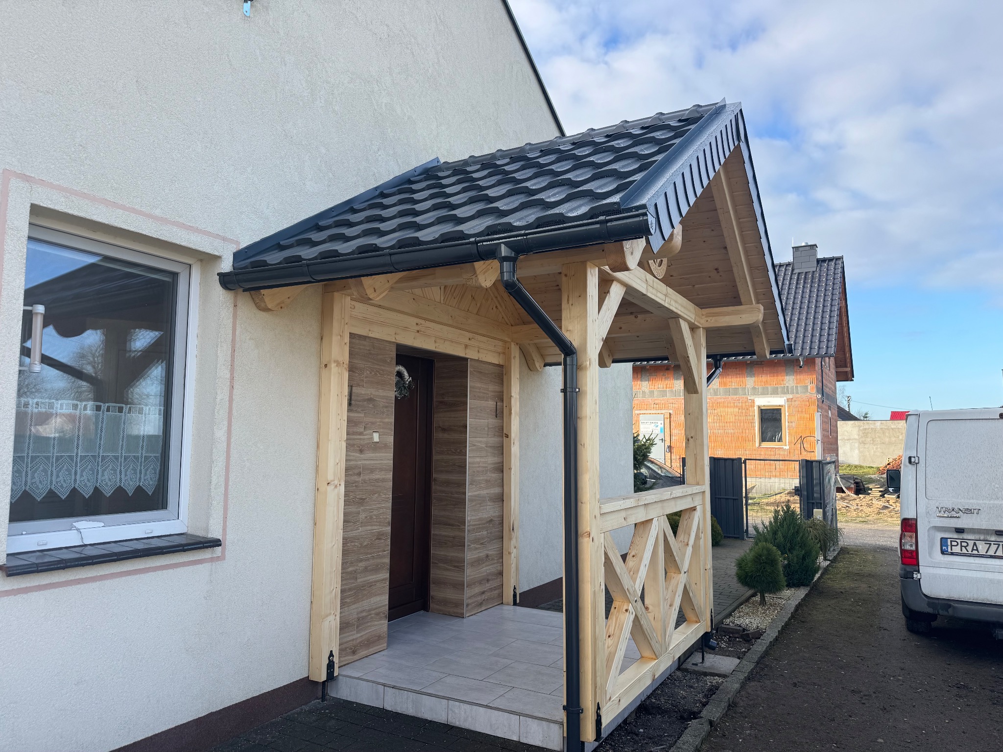 Drewniana wiata nad wejściem do domu z czarnym dachem i rynnami. Widoczna elewacja, okno i fragment podjazdu. Nowoczesny design z elementami dekoracyjnymi.