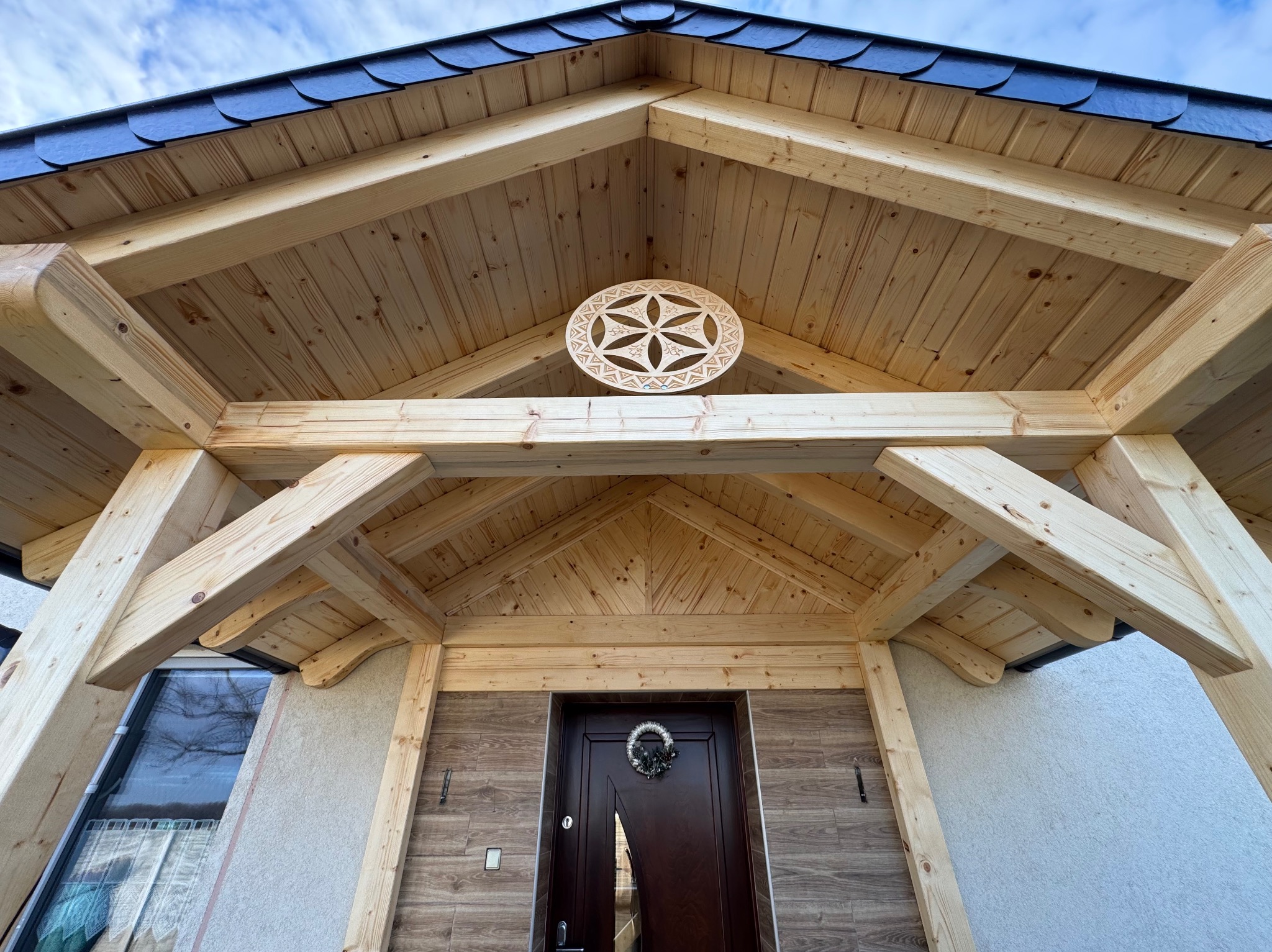 Drewniana konstrukcja z elementami dekoracyjnymi nad wejściem do domu, widok z dołu. Ciemny dach z łupka. Na suficie ozdobny, ażurowy element.