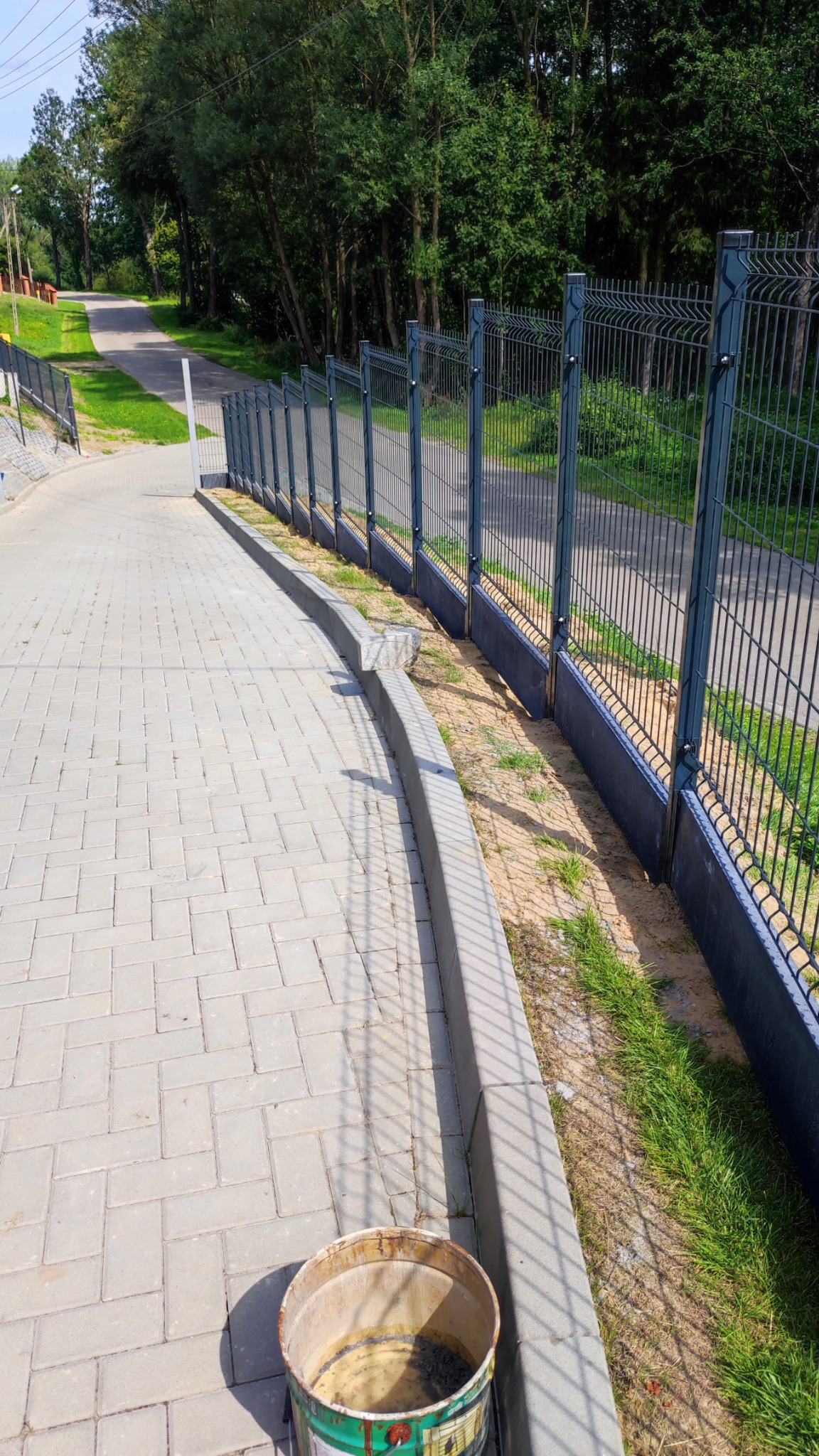 Długa, metalowa siatka ogrodzeniowa wzdłuż chodnika z kostki, z widocznym wiadrem z resztkami zaprawy. Tło: drzewa i droga.