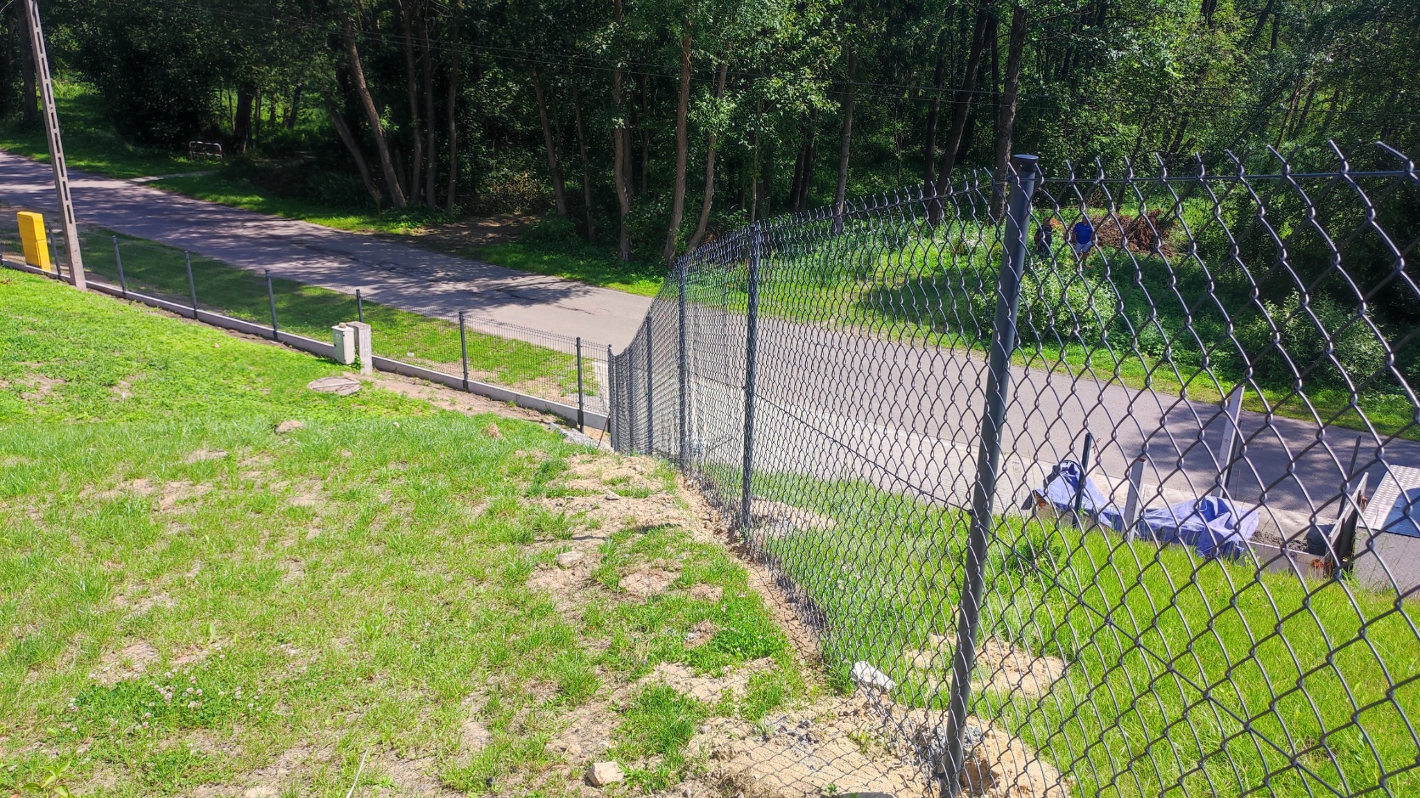Długa siatka ogrodzeniowa z betonową podmurówką, biegnąca wzdłuż drogi, na tle zieleni. Widoczne słupki i naprężenia siatki. Drobne nierówności terenu.