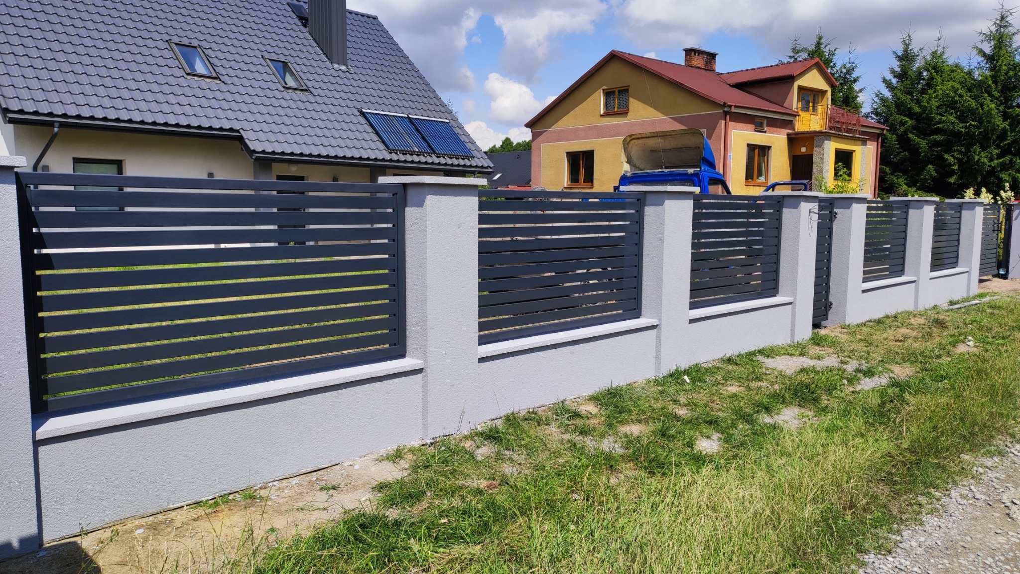 Nowoczesne ogrodzenie metalowe w kolorze grafitowym z podmurówką, w tle domy jednorodzinne i błękitne niebo.