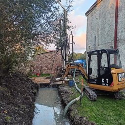AMBROCH SPÓŁKA Z OGRANICZONĄ ODPOWIEDZIALNOŚCIĄ - Wiercenie studni głębinowej przy użyciu wiertnicy i minikoparki CAT 301.7D. W tle budynek z cegły i otynkowany. W wykopie woda. Prace w toku.
