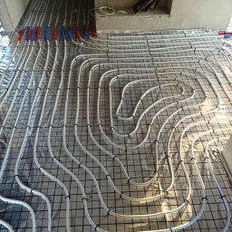 AMBROCH SPÓŁKA Z OGRANICZONĄ ODPOWIEDZIALNOŚCIĄ - Ułożone rury ogrzewania podłogowego na folii, przymocowane do siatki zbrojeniowej. Widoczne fragmenty rur w kolorze niebieskim i czerwonym. Ujęcie z góry, w tle schody.