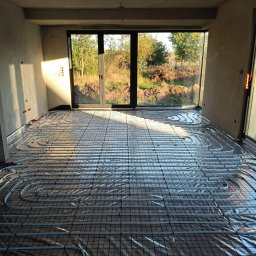 AMBROCH SPÓŁKA Z OGRANICZONĄ ODPOWIEDZIALNOŚCIĄ - Instalacja ogrzewania podłogowego w surowym wnętrzu z dużym oknem. Widoczne rury grzewcze ułożone na folii i siatce, przygotowane do zalania posadzką. Surowe ściany i naturalne światło.