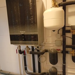 AMBROCH SPÓŁKA Z OGRANICZONĄ ODPOWIEDZIALNOŚCIĄ - Zamontowany, wiszący piec gazowy Buderus z podłączonymi rurami i osprzętem na tle białej ściany z płytkami. Widoczne zawory, manometr i zbiornik wyrównawczy.