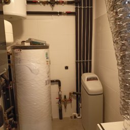 AMBROCH SPÓŁKA Z OGRANICZONĄ ODPOWIEDZIALNOŚCIĄ - Biały zasobnik ciepłej wody owinięty folią, obok zmiękczacz wody i rury w izolacji na tle jasnych płytek. Widoczne zawory i manometry.