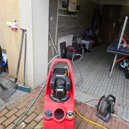 AMBROCH SPÓŁKA Z OGRANICZONĄ ODPOWIEDZIALNOŚCIĄ - Czerwona pompa do instalacji grzewczych Sentinel na kostce brukowej, z widocznymi przewodami i elementami instalacji w tle garażu. Sprzęt do czyszczenia instalacji.