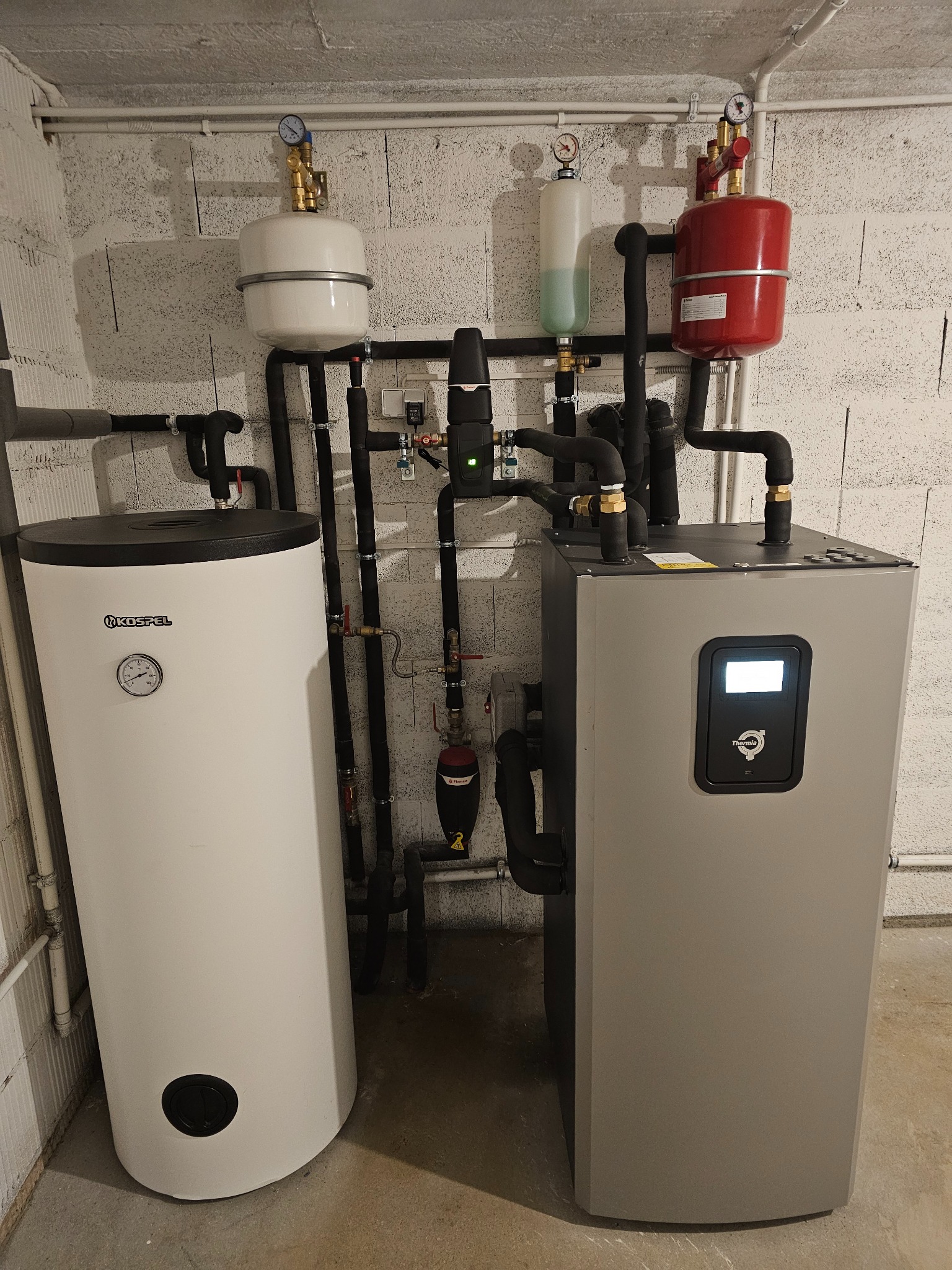 Biały zbiornik Kospel i szary piec Thermia z czarnymi rurami w kotłowni z betonową ścianą i podłogą. Widoczne manometry i zawory.