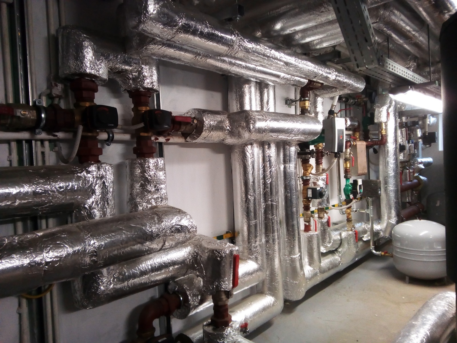 Izolowane rury instalacji grzewczej w kotłowni, z widocznymi zaworami i elementami sterującymi. Rury owinięte srebrną folią izolacyjną, tworzące labirynt instalacji.