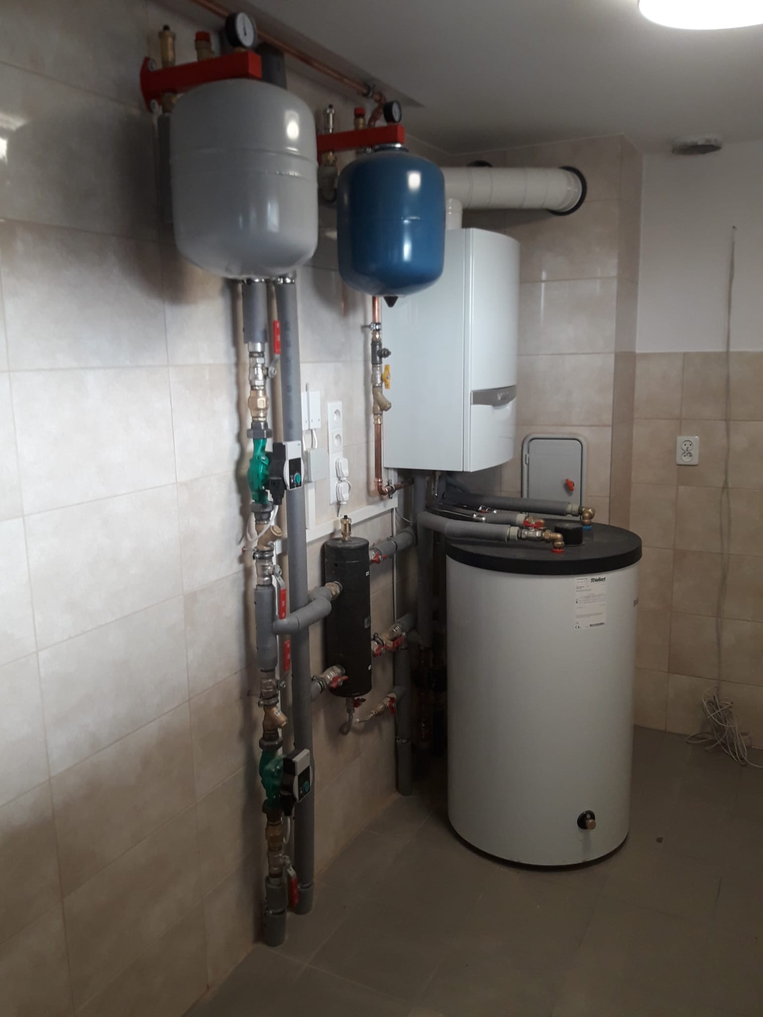 Zestaw instalacji grzewczej z kotłem gazowym, zbiornikiem i naczyniami wyrównawczymi w kotłowni wykończonej płytkami. Widoczne rury, zawory i manometr.