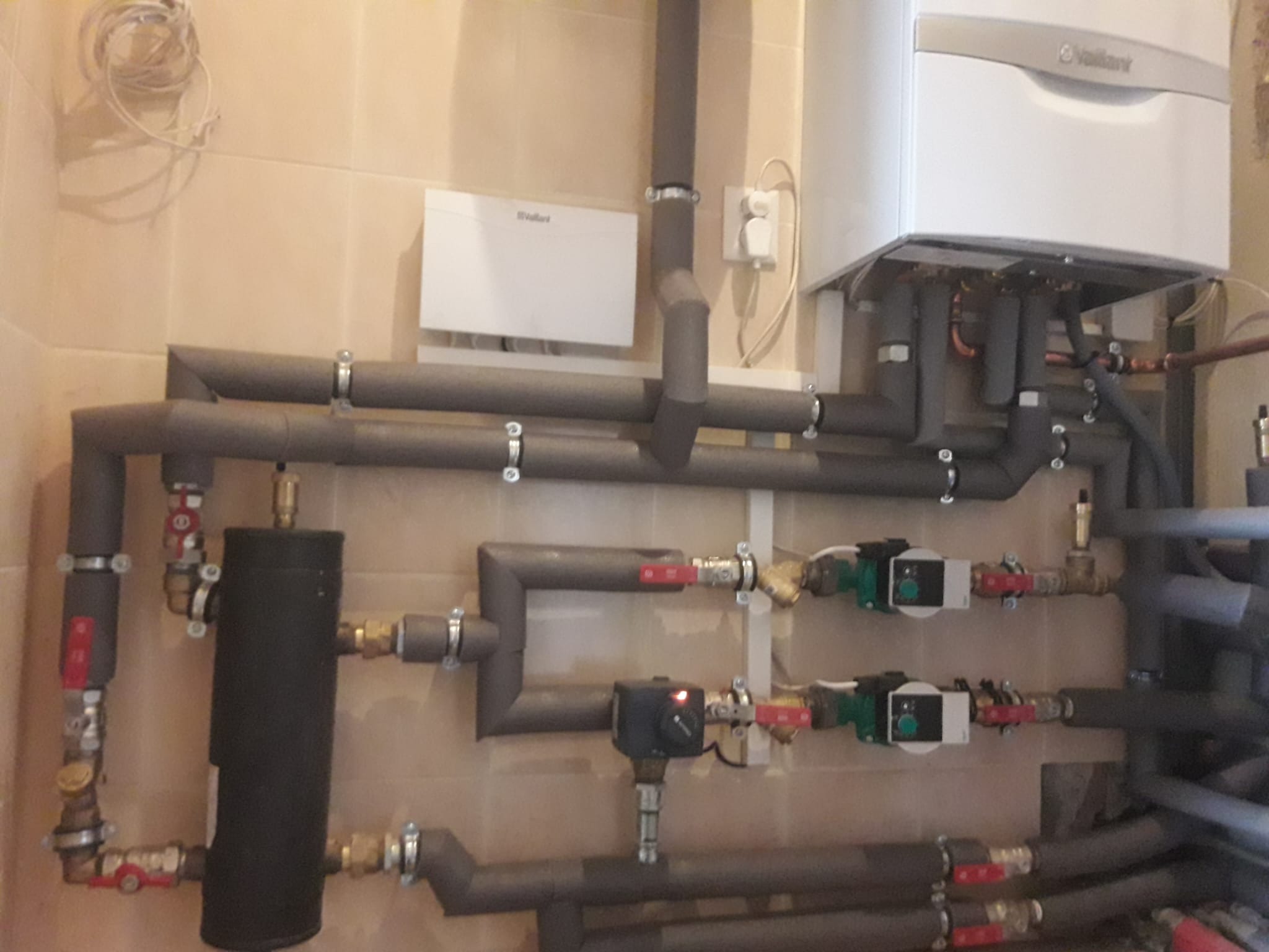 Złożona instalacja grzewcza z kotłem Vaillant, rurami w izolacji, pompami obiegowymi i zaworami, zamontowana na beżowych płytkach. Widoczne elementy sterujące i zasilanie.