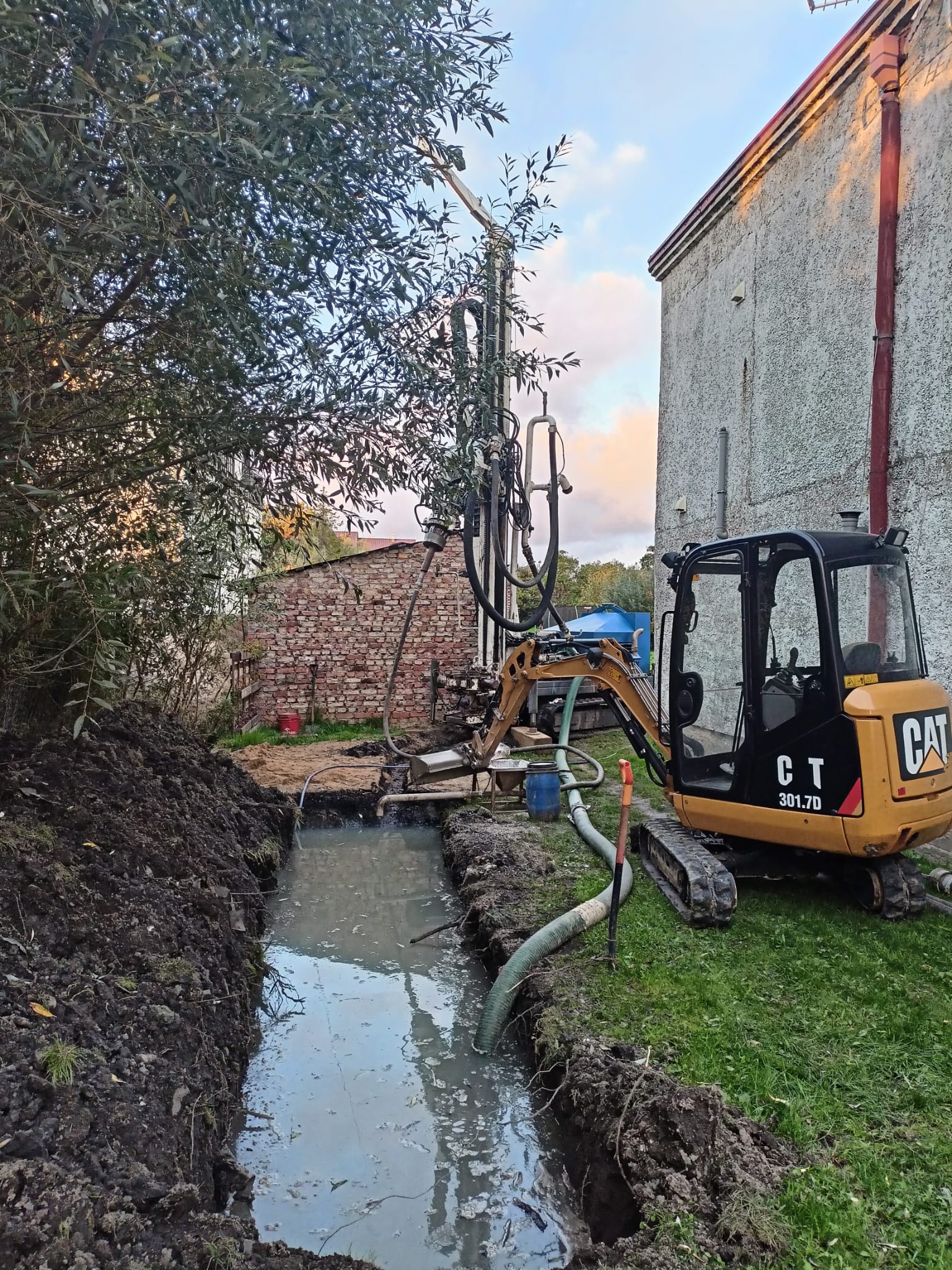 Wiercenie studni głębinowej przy użyciu wiertnicy i minikoparki CAT 301.7D. W tle budynek z cegły i otynkowany. W wykopie woda. Prace w toku.