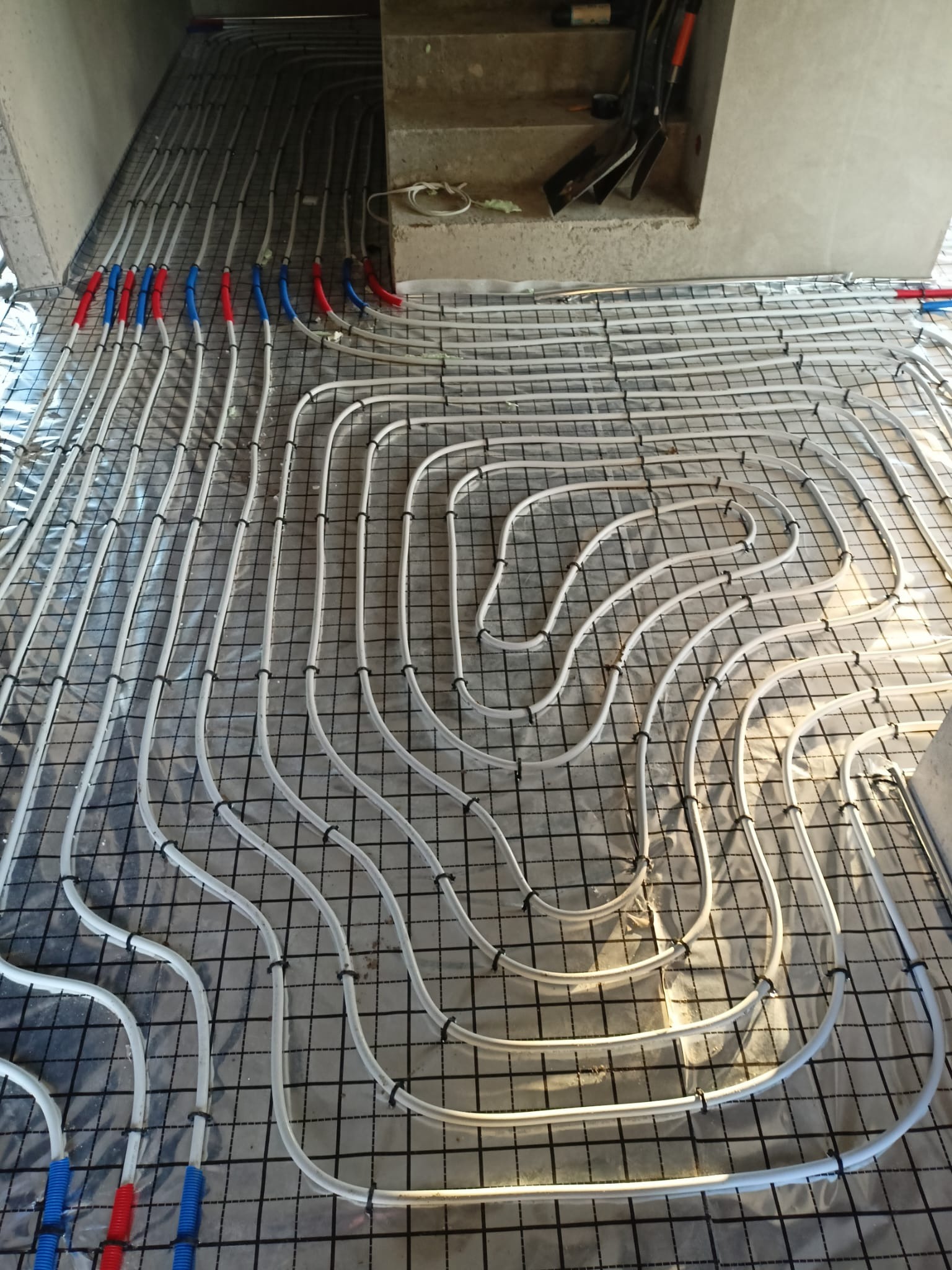 Ułożone rury ogrzewania podłogowego na folii, przymocowane do siatki zbrojeniowej. Widoczne fragmenty rur w kolorze niebieskim i czerwonym. Ujęcie z góry, w tle schody.