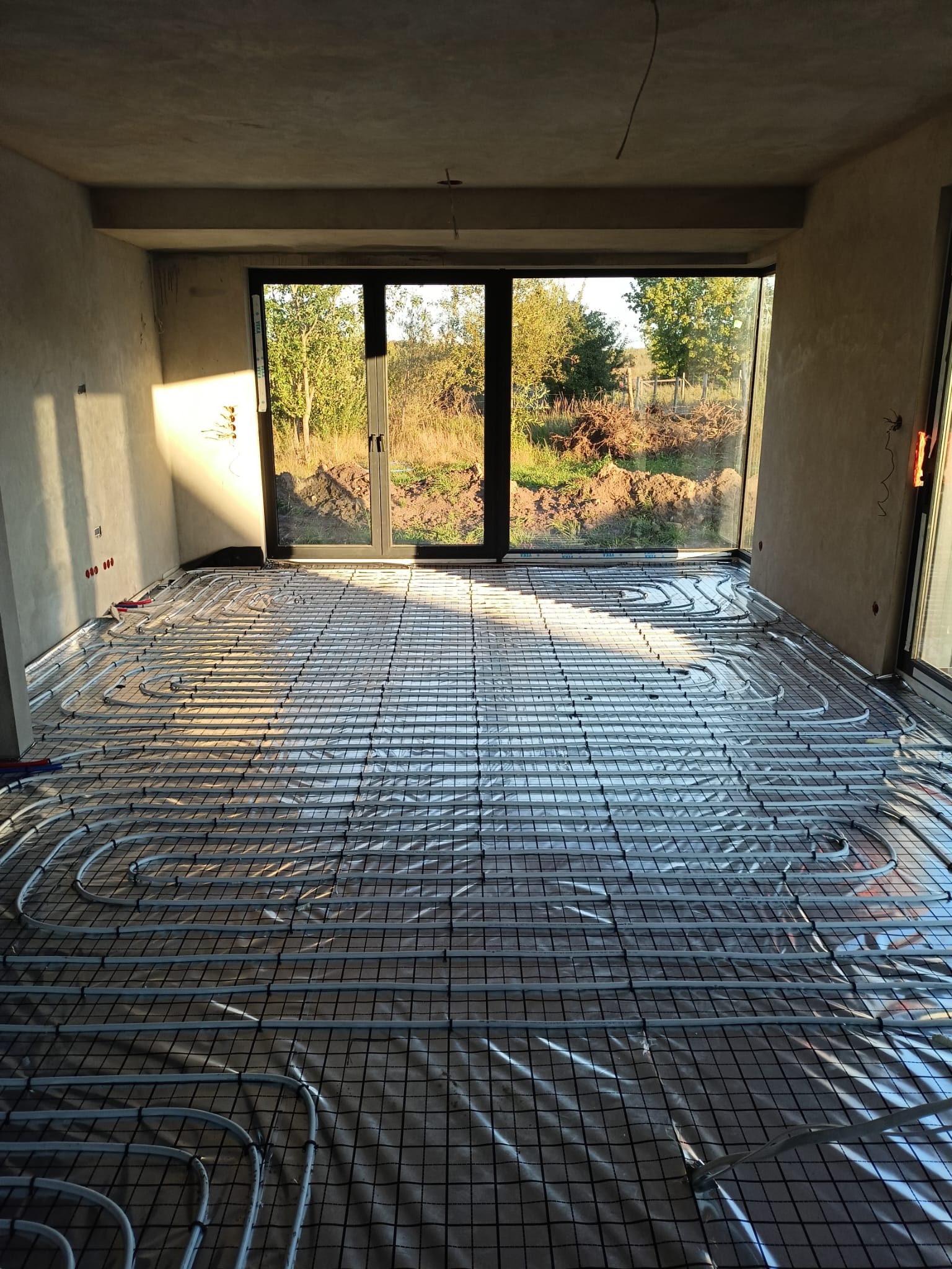 Instalacja ogrzewania podłogowego w surowym wnętrzu z dużym oknem. Widoczne rury grzewcze ułożone na folii i siatce, przygotowane do zalania posadzką. Surowe ściany i naturalne światło.