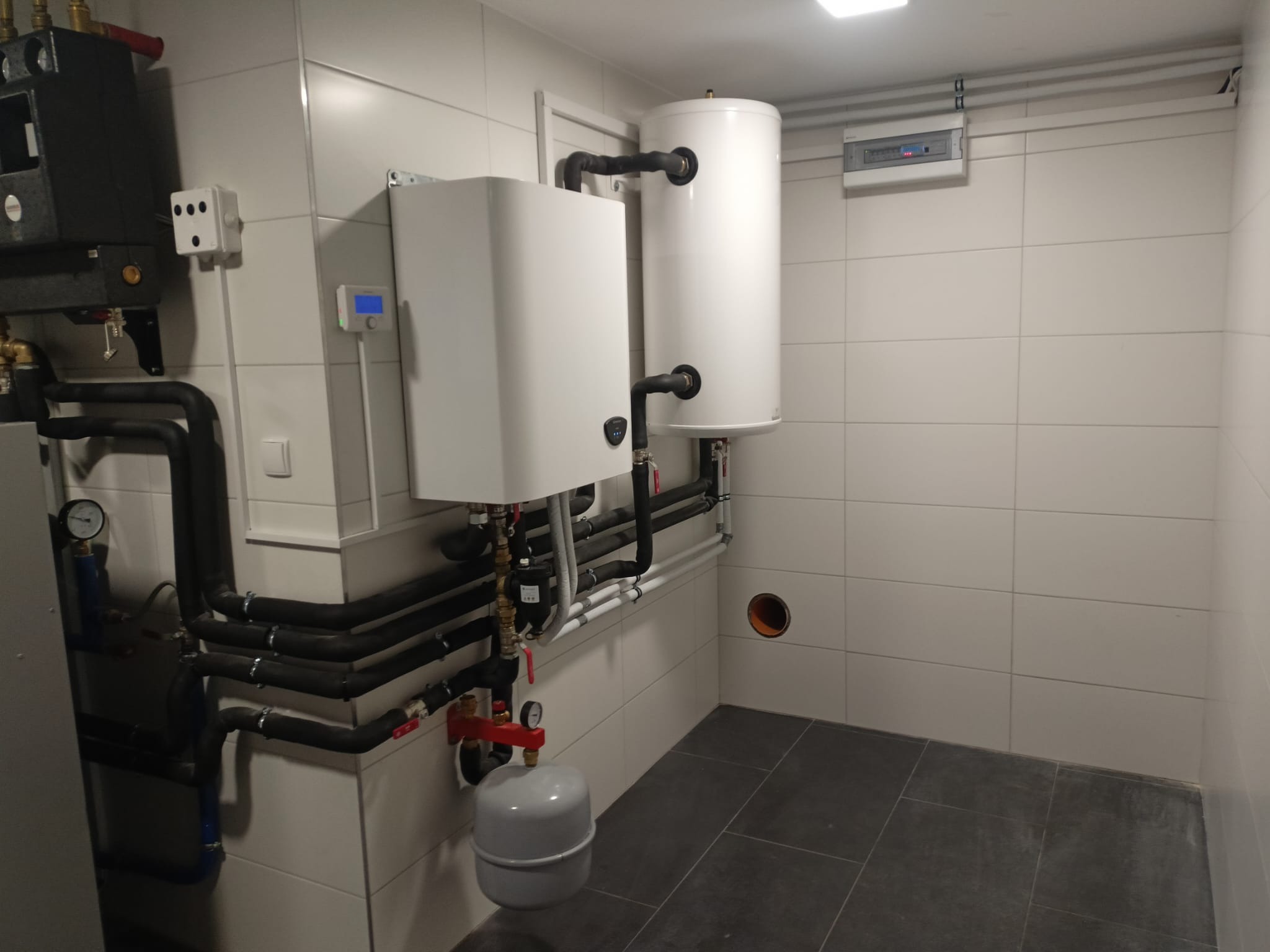 Białe kotły grzewcze i czarne rury w schludnej kotłowni wyłożonej białymi płytkami. Szare płytki na podłodze. Widoczna instalacja elektryczna.