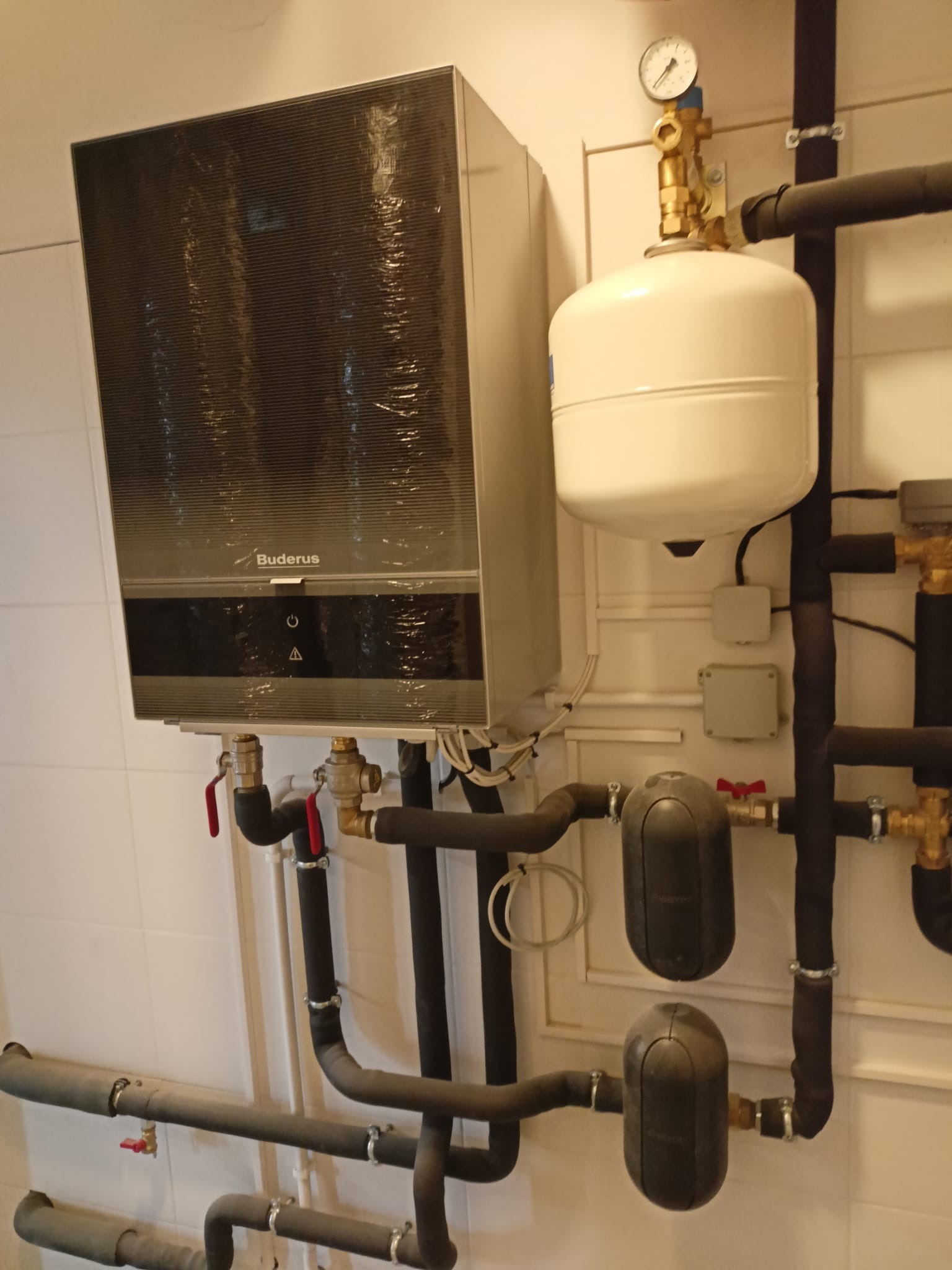 Zamontowany, wiszący piec gazowy Buderus z podłączonymi rurami i osprzętem na tle białej ściany z płytkami. Widoczne zawory, manometr i zbiornik wyrównawczy.