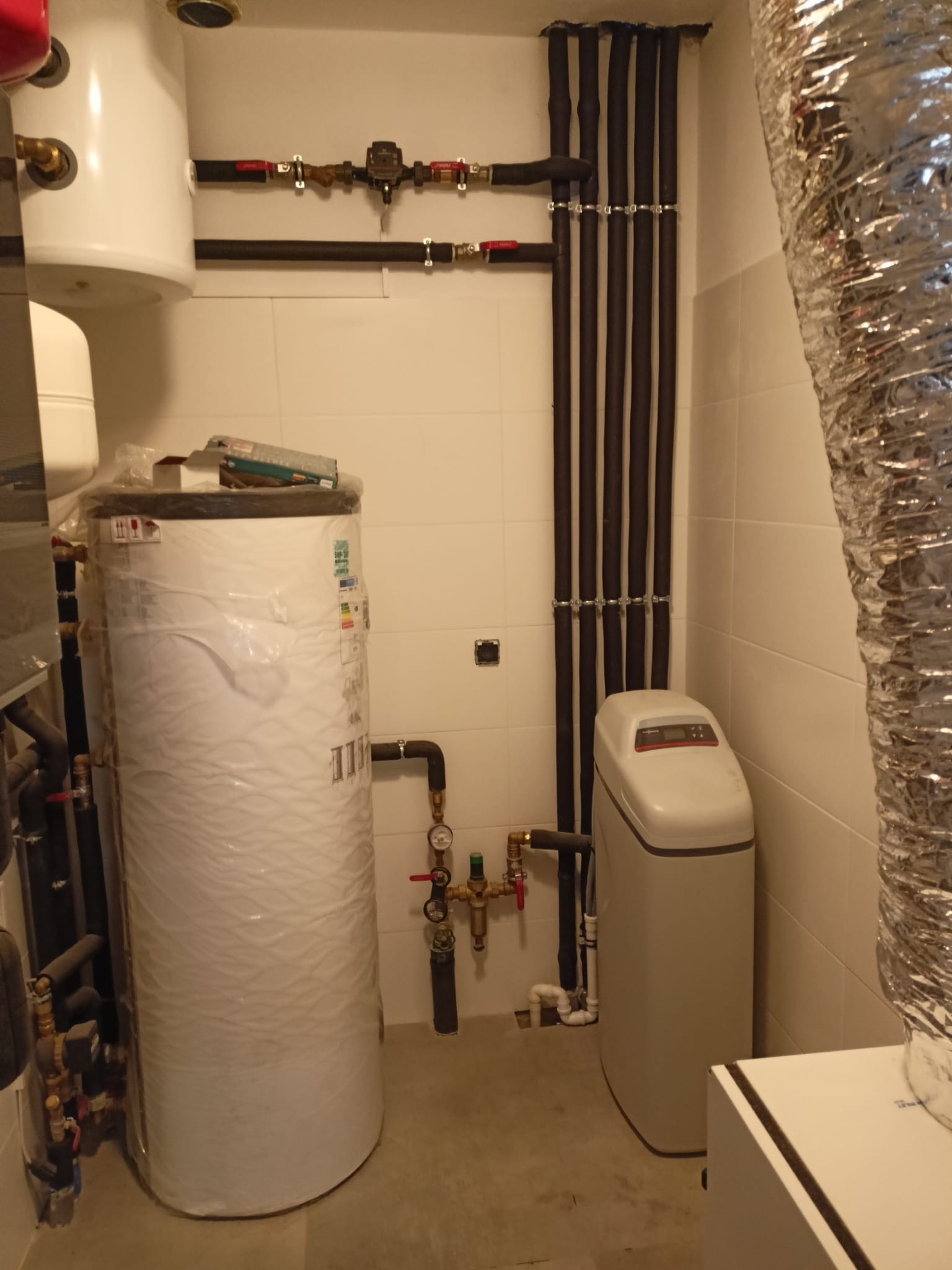 Biały zasobnik ciepłej wody owinięty folią, obok zmiękczacz wody i rury w izolacji na tle jasnych płytek. Widoczne zawory i manometry.