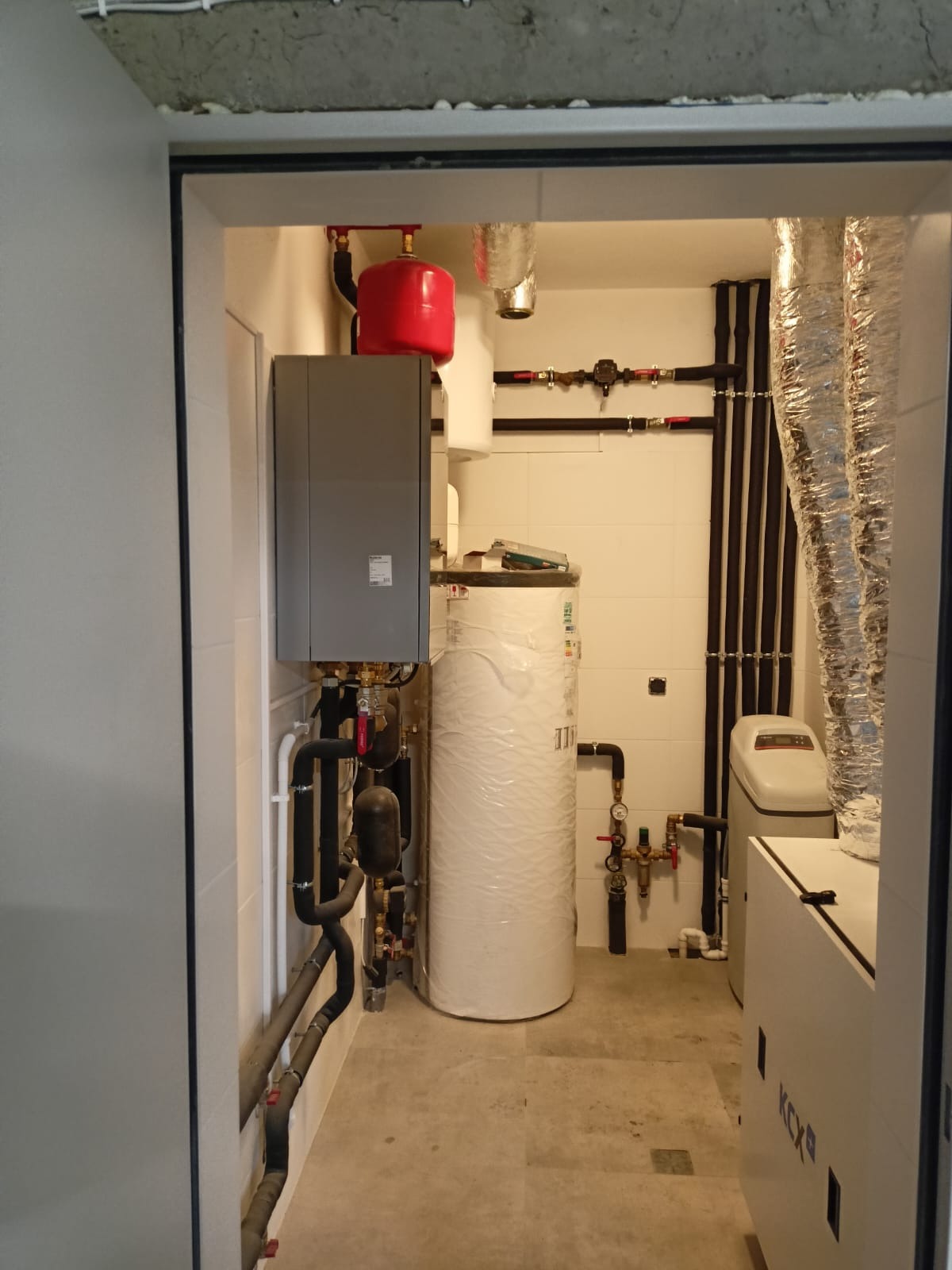 Kompletna instalacja grzewcza w kotłowni: szary piec, biały zbiornik owinięty folią, czerwony zbiornik wyrównawczy, czarne rury i biały zmiękczacz wody. Widok przez futrynę.
