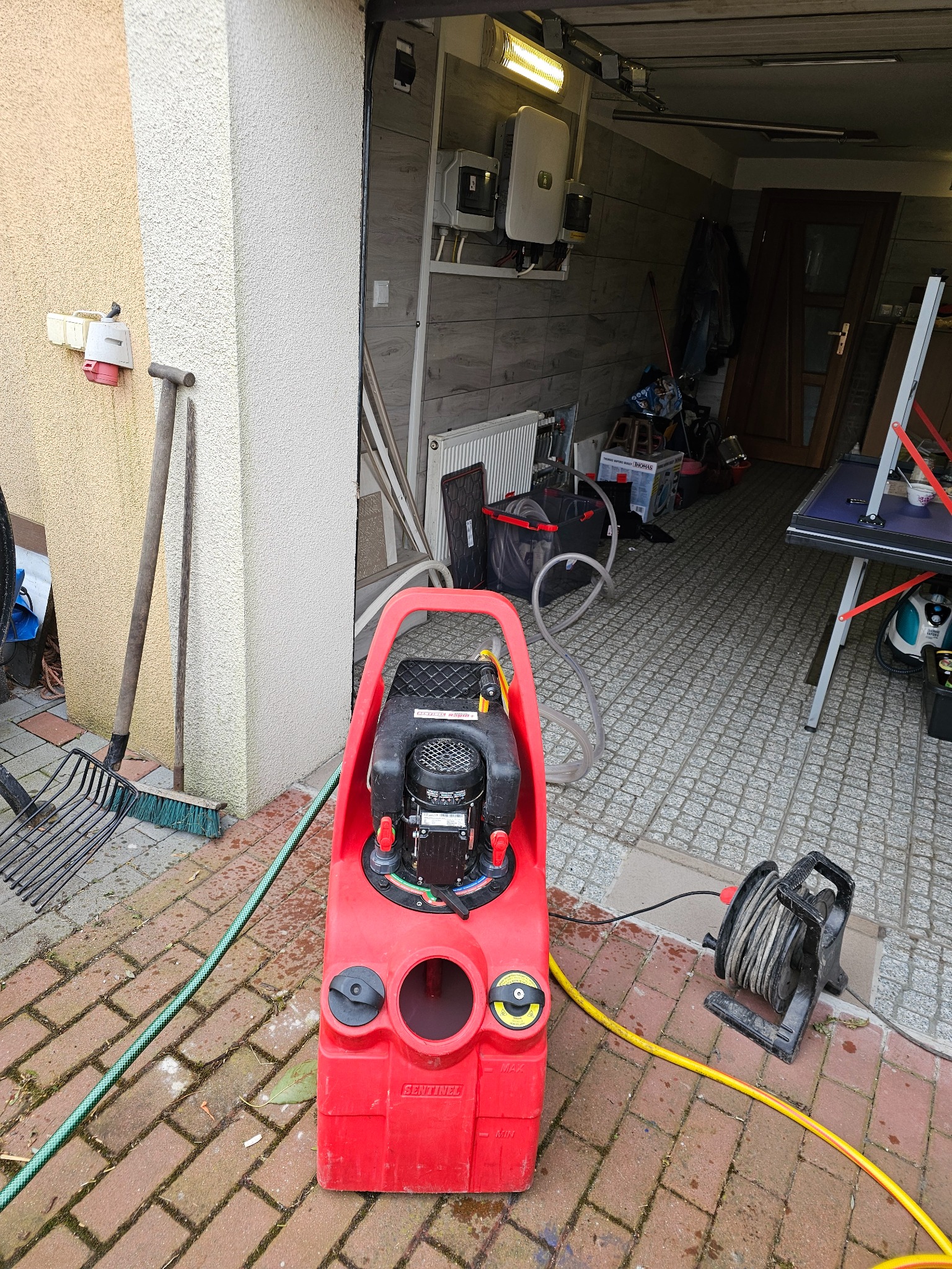 Czerwona pompa do instalacji grzewczych Sentinel na kostce brukowej, z widocznymi przewodami i elementami instalacji w tle garażu. Sprzęt do czyszczenia instalacji.