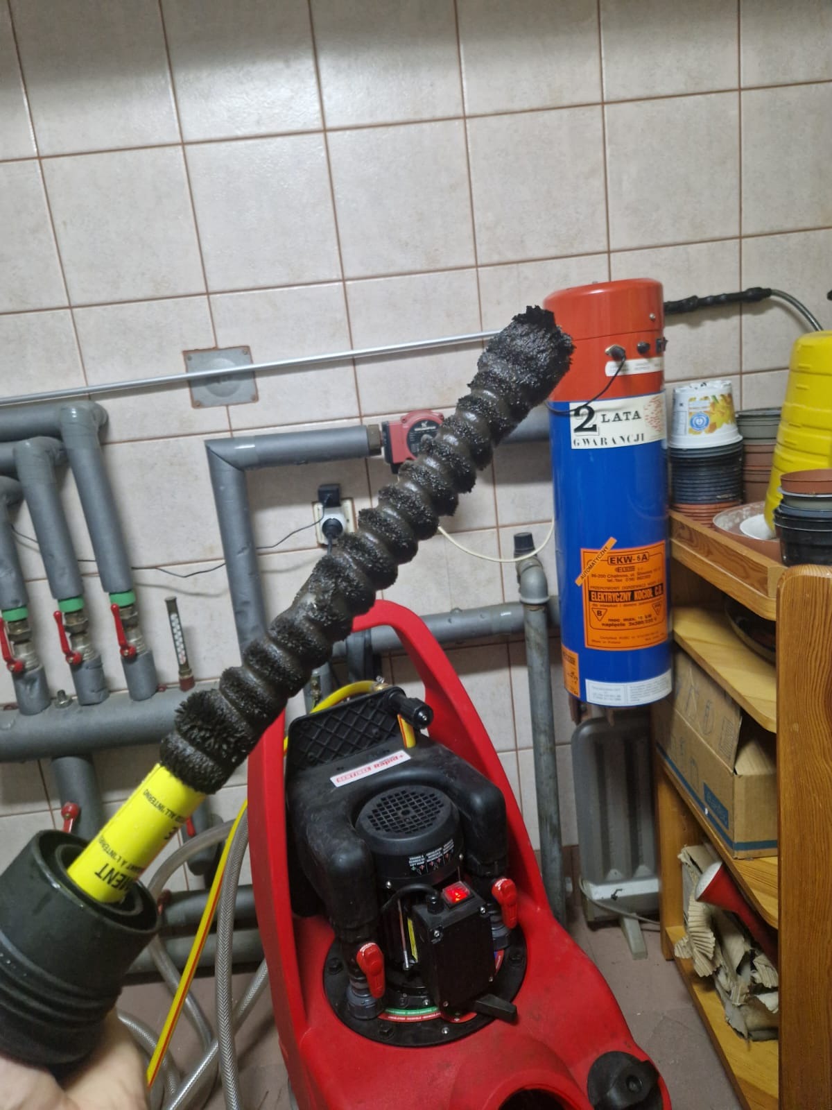 Czyszczenie instalacji grzewczej: pompa w czerwonej obudowie z podłączonym wężem, widoczne rury instalacji i bojler elektryczny w tle.