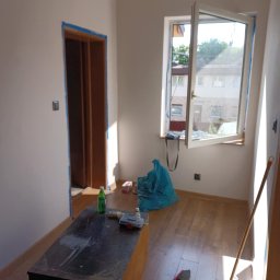 ProMax - Pokój w trakcie przygotowań do malowania: zabezpieczone taśmą okno i futryna, widoczne narzędzia, podłoga częściowo zakryta. Przygotowanie do malowania ścian.