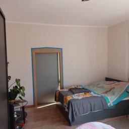 ProMax - Pokój przed odświeżeniem: ściany w neutralnym odcieniu, zabezpieczone taśmą malarską framugi drzwi, drewniana podłoga i widoczne meble.