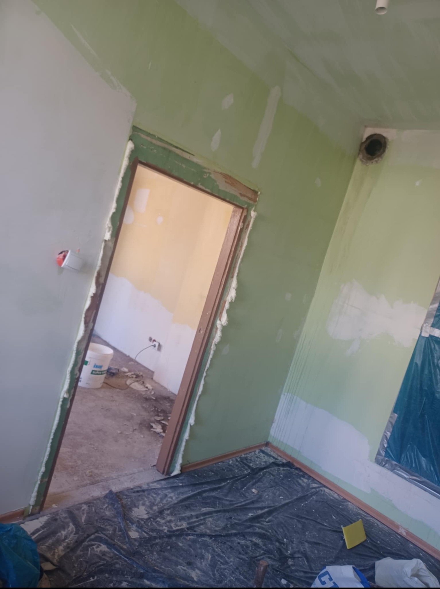 Pokój w trakcie prac wykończeniowych: ściany częściowo pomalowane na zielono, fragmenty szpachli, podłoga zabezpieczona folią. Widoczny otwór wentylacyjny w suficie.