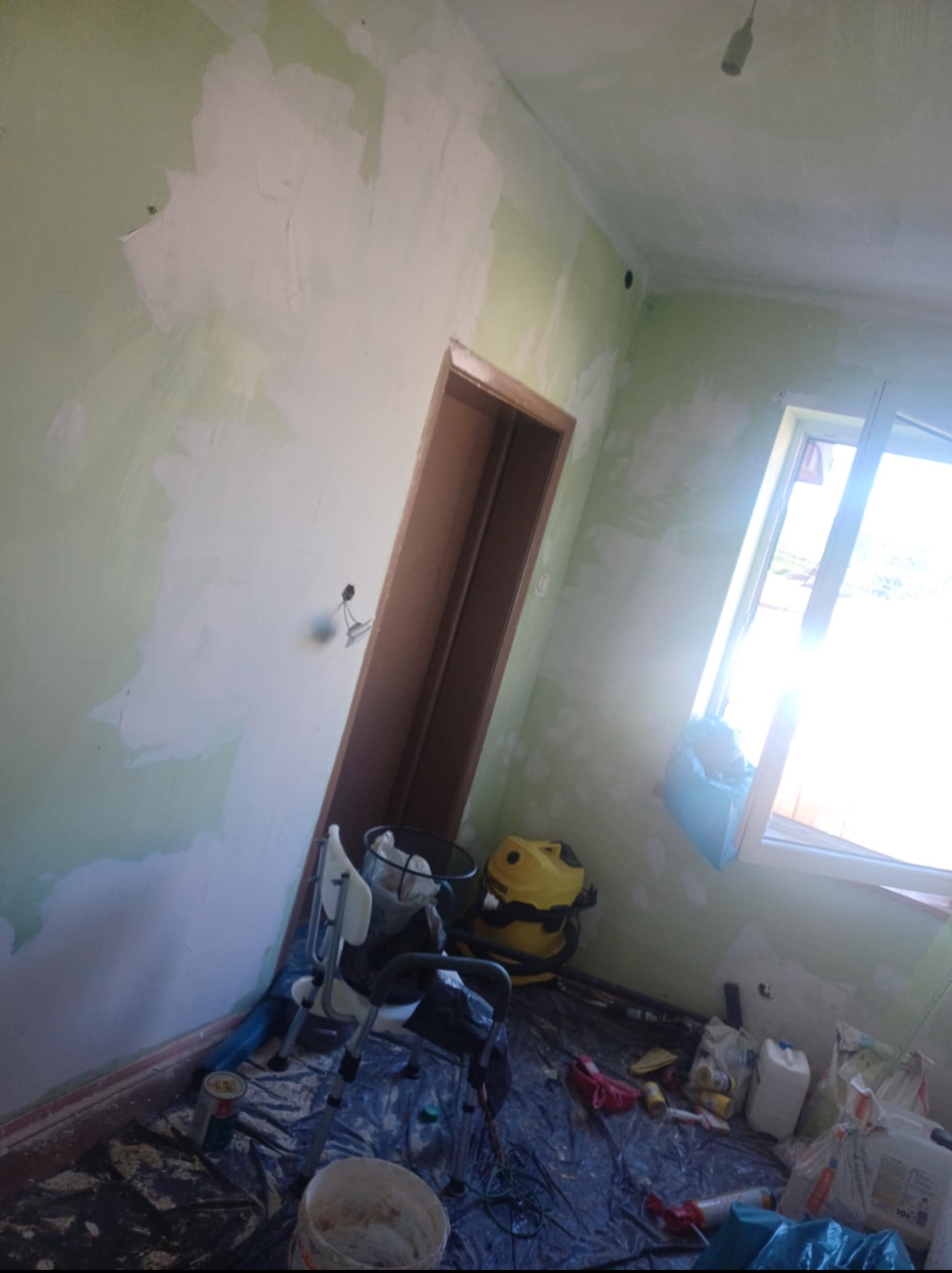 Pokój w trakcie szpachlowania: ściany z nałożoną gładzią, widoczne narzędzia, wiadro z masą, folia ochronna na podłodze, okno z intensywnym światłem.