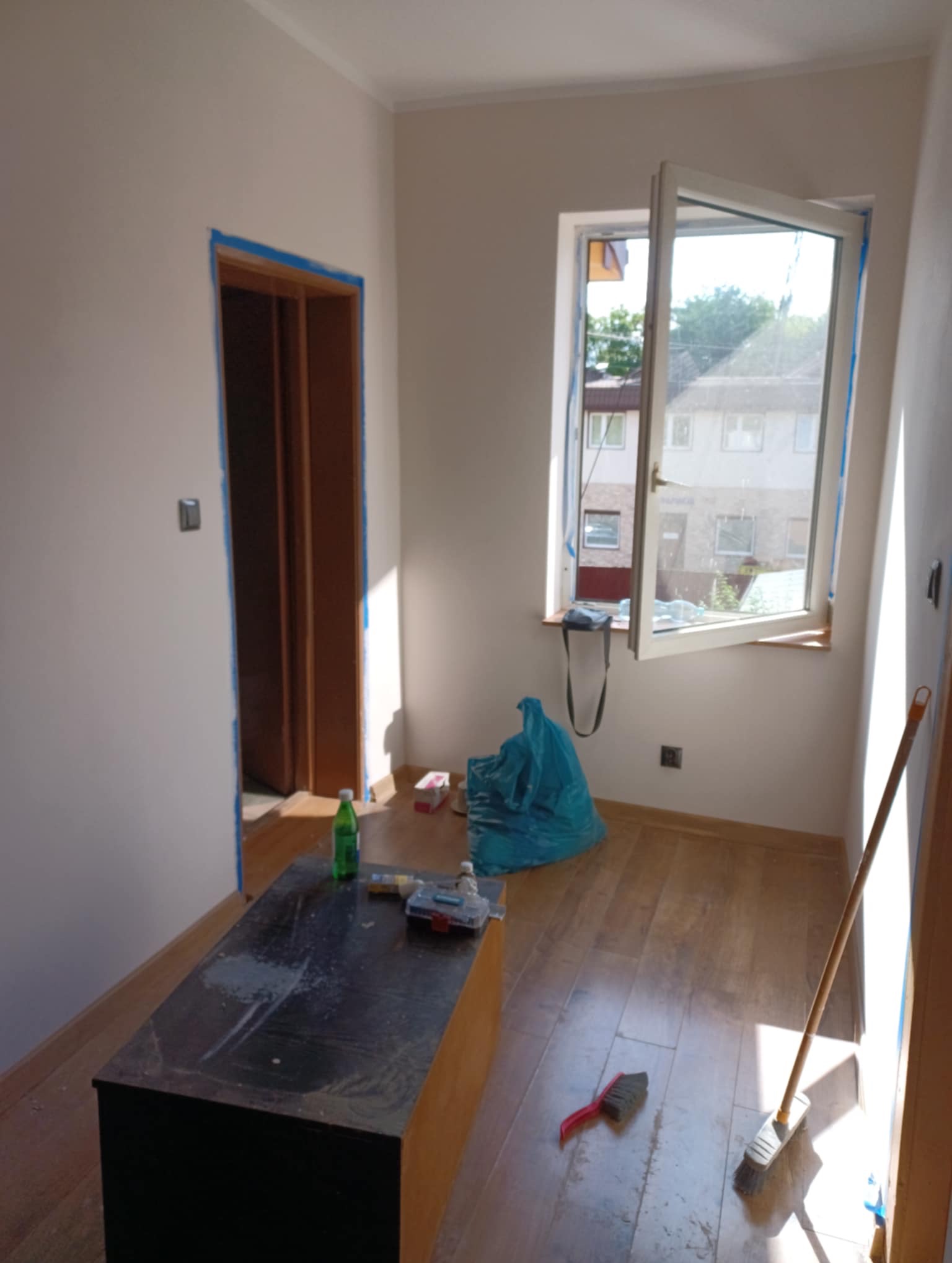 Pokój w trakcie przygotowań do malowania: zabezpieczone taśmą okno i futryna, widoczne narzędzia, podłoga częściowo zakryta. Przygotowanie do malowania ścian.
