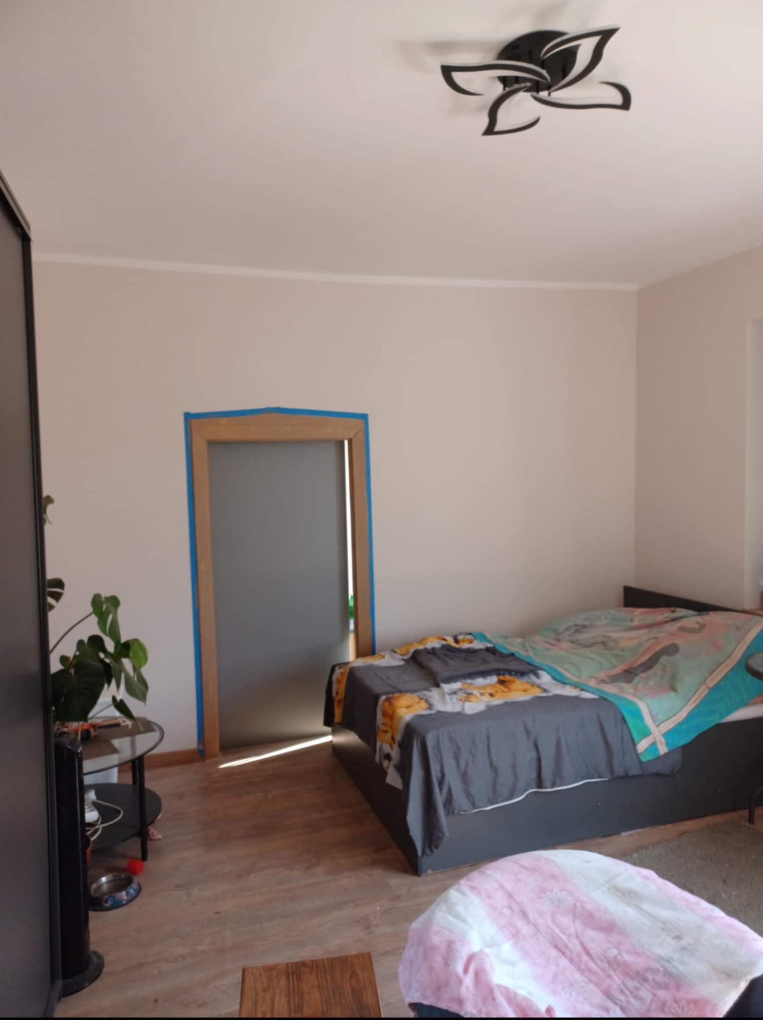 Pokój przed odświeżeniem: ściany w neutralnym odcieniu, zabezpieczone taśmą malarską framugi drzwi, drewniana podłoga i widoczne meble.