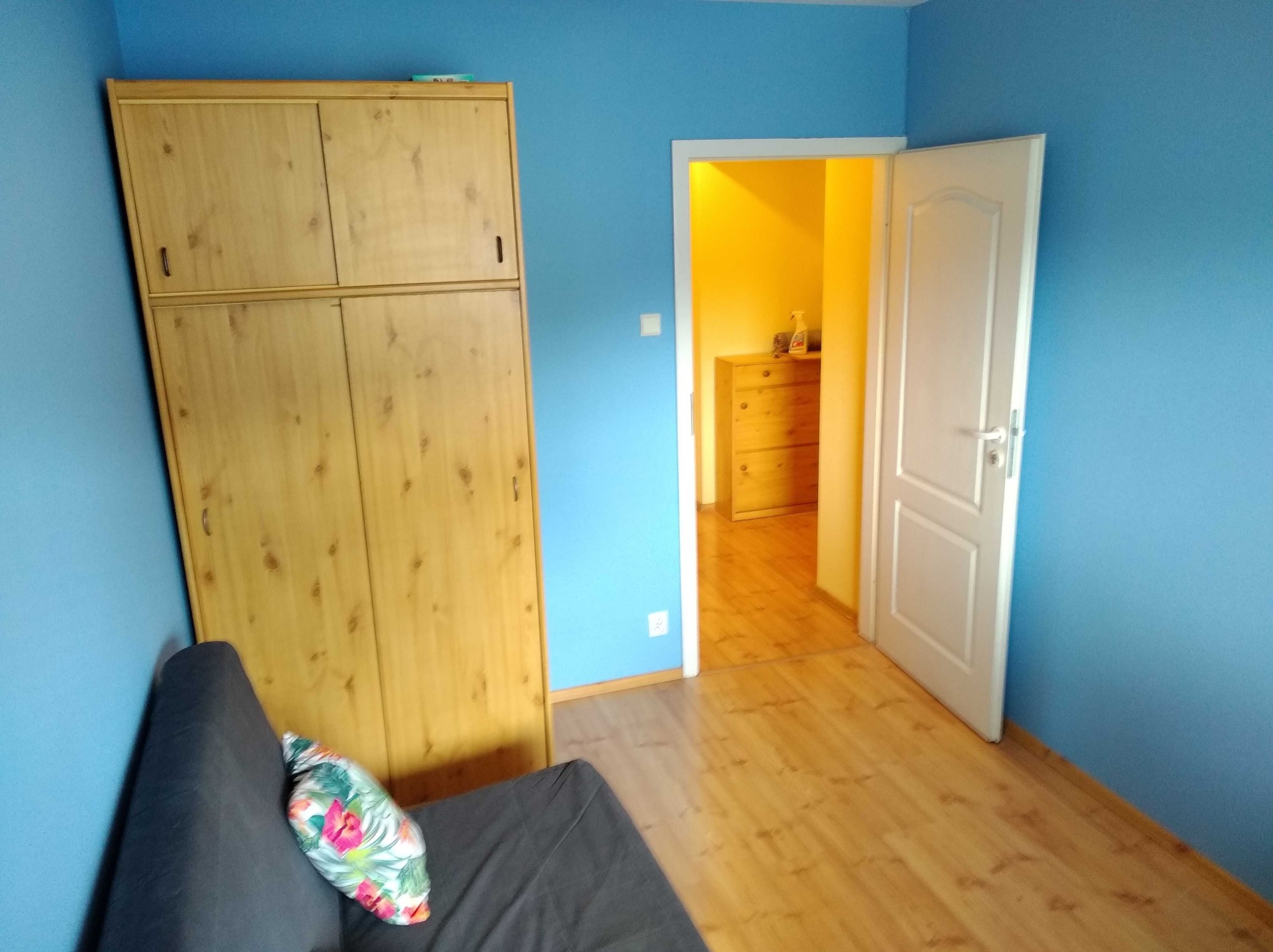 Błękitny pokój z drewnianą szafą, kanapą i otwartymi drzwiami do żółtego pomieszczenia. Widoczne panele podłogowe i dekoracyjna poduszka na kanapie.