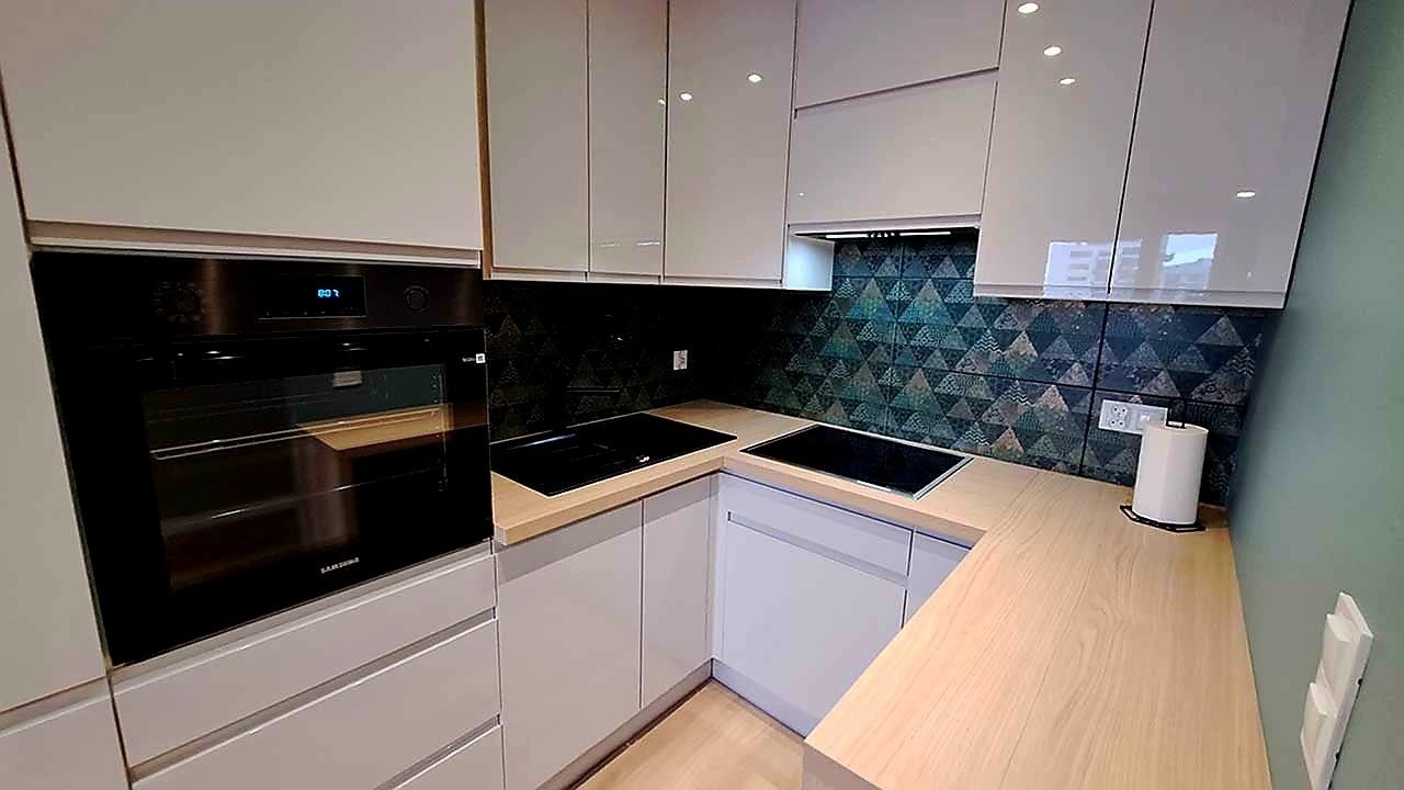 Nowoczesna kuchnia z białymi szafkami, drewnianym blatem i mozaiką na ścianie. Widoczny piekarnik Samsung i płyta indukcyjna. Minimalistyczny design.