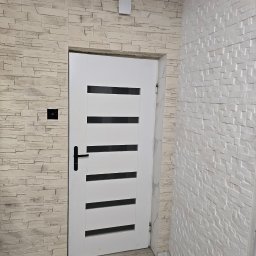 Remonty i wykończenia Marcin Dydak - Białe drzwi z czarną klamką w Wojkowicach Kościelnych, wkomponowane w ścianę wyłożoną dekoracyjną płytką imitującą kamień. Nowoczesny design.