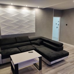 Remonty i wykończenia Marcin Dydak - Szary salon z narożnikiem i dekoracyjną falistą płytą ścienną. Ściany wykończone w stylu betonu architektonicznego. Widoczne oświetlenie punktowe i minimalistyczny stolik.