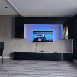 Remonty i wykończenia Marcin Dydak - Nowoczesny salon z podświetlonym telewizorem, czarnymi meblami i szarą ścianą. Widoczne krzesła i panele podłogowe. Oświetlenie LED za telewizorem.