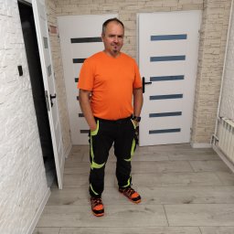 Remonty i wykończenia Marcin Dydak - Mężczyzna w stroju roboczym stoi na tle wykończonego wnętrza z dwiema białymi, nowoczesnymi drzwiami i ścianą z dekoracyjnej cegły. Widoczna grzejnik i podłoga.