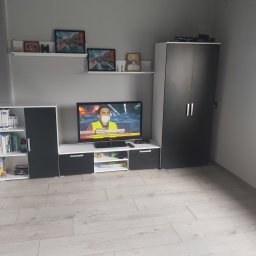 Remonty i wykończenia Marcin Dydak - Nowoczesny salon w odcieniach szarości i bieli. Meble w zabudowie, telewizor, dekoracje ścienne. Jasna podłoga z paneli. Minimalistyczny design.